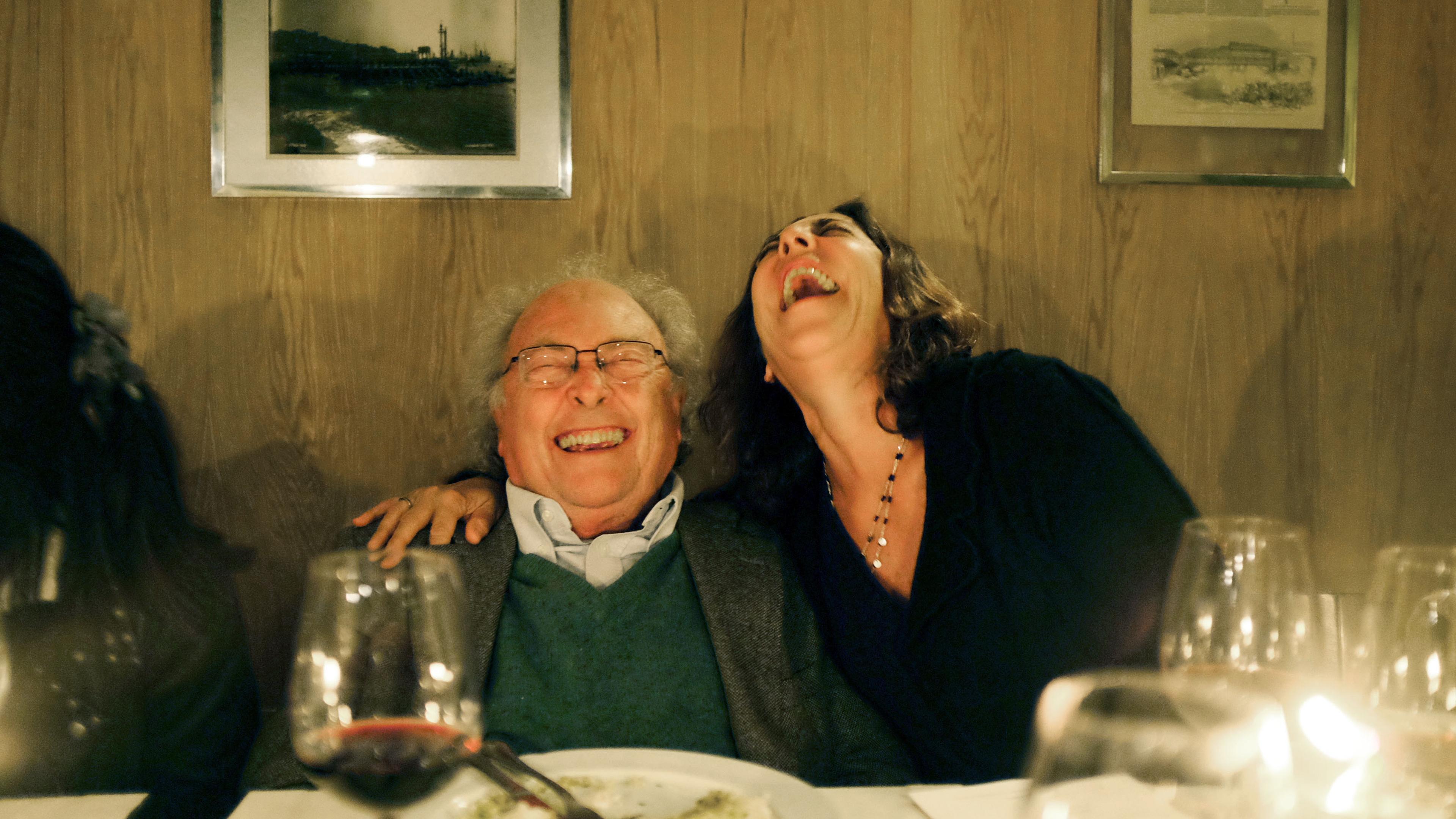 Photo of two people at a dinner table laughing heartily with wine glasses and a plate in the foreground.