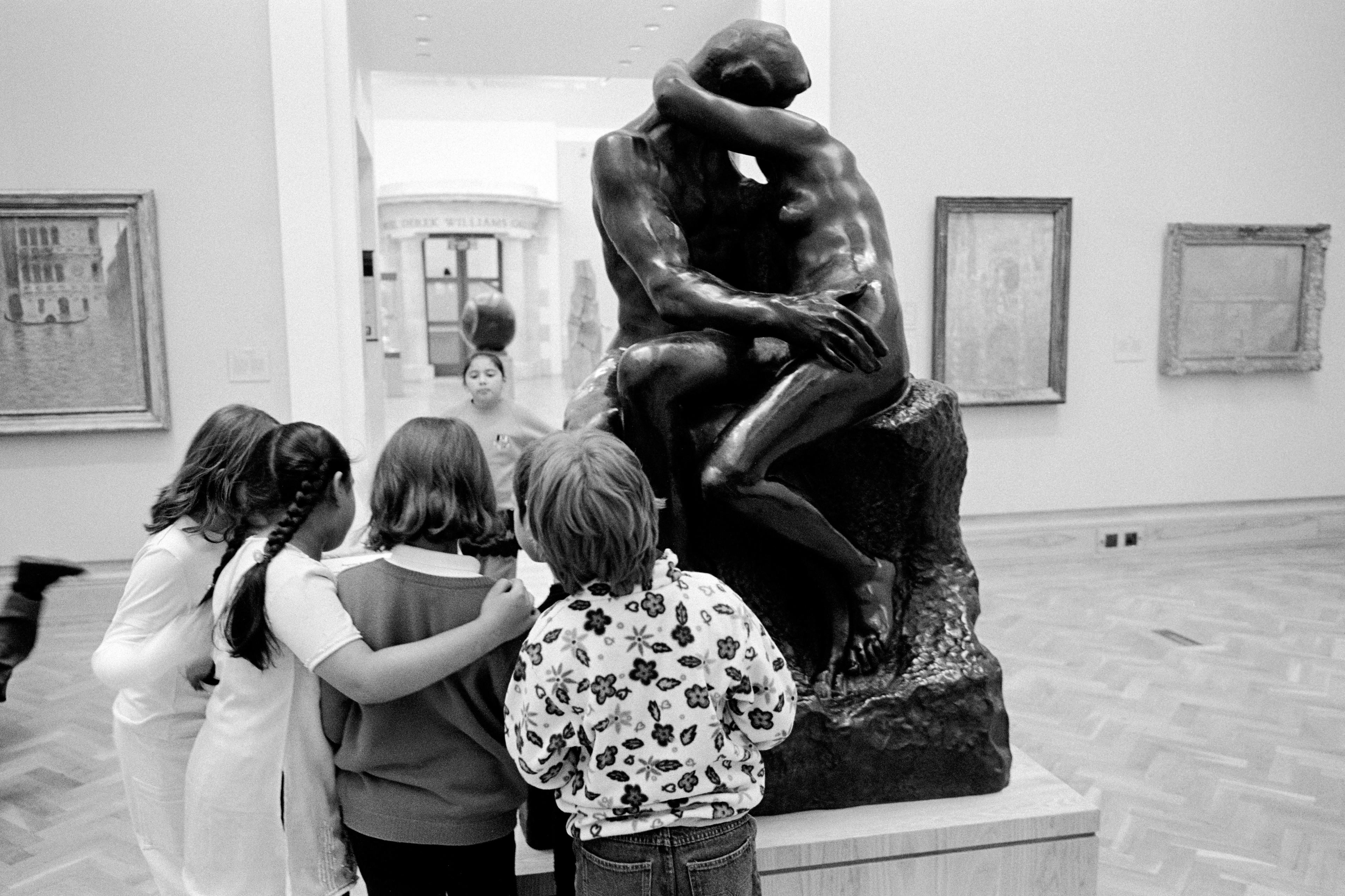 Photo of children observing a large sculpture of a couple embracing in a museum setting with paintings in the background.