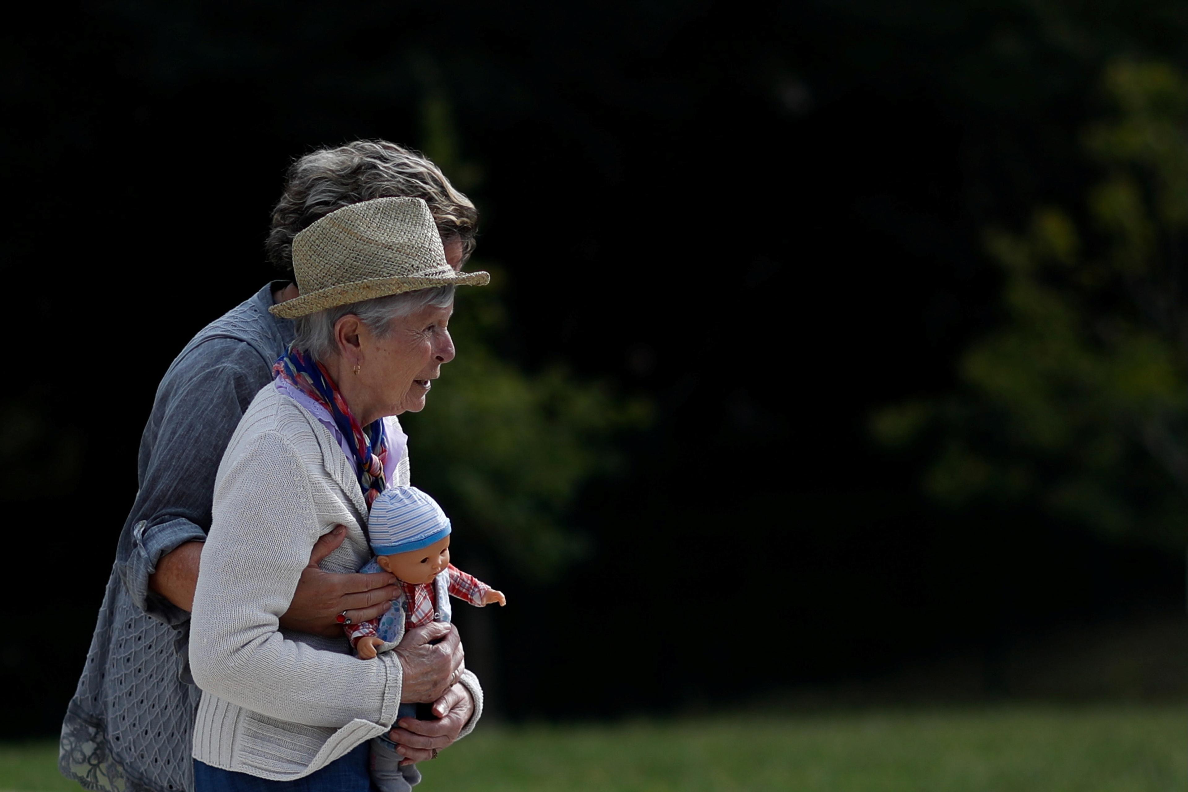 Photo of an older woman in a straw hat holding a doll in a park with another person embracing her from behind.