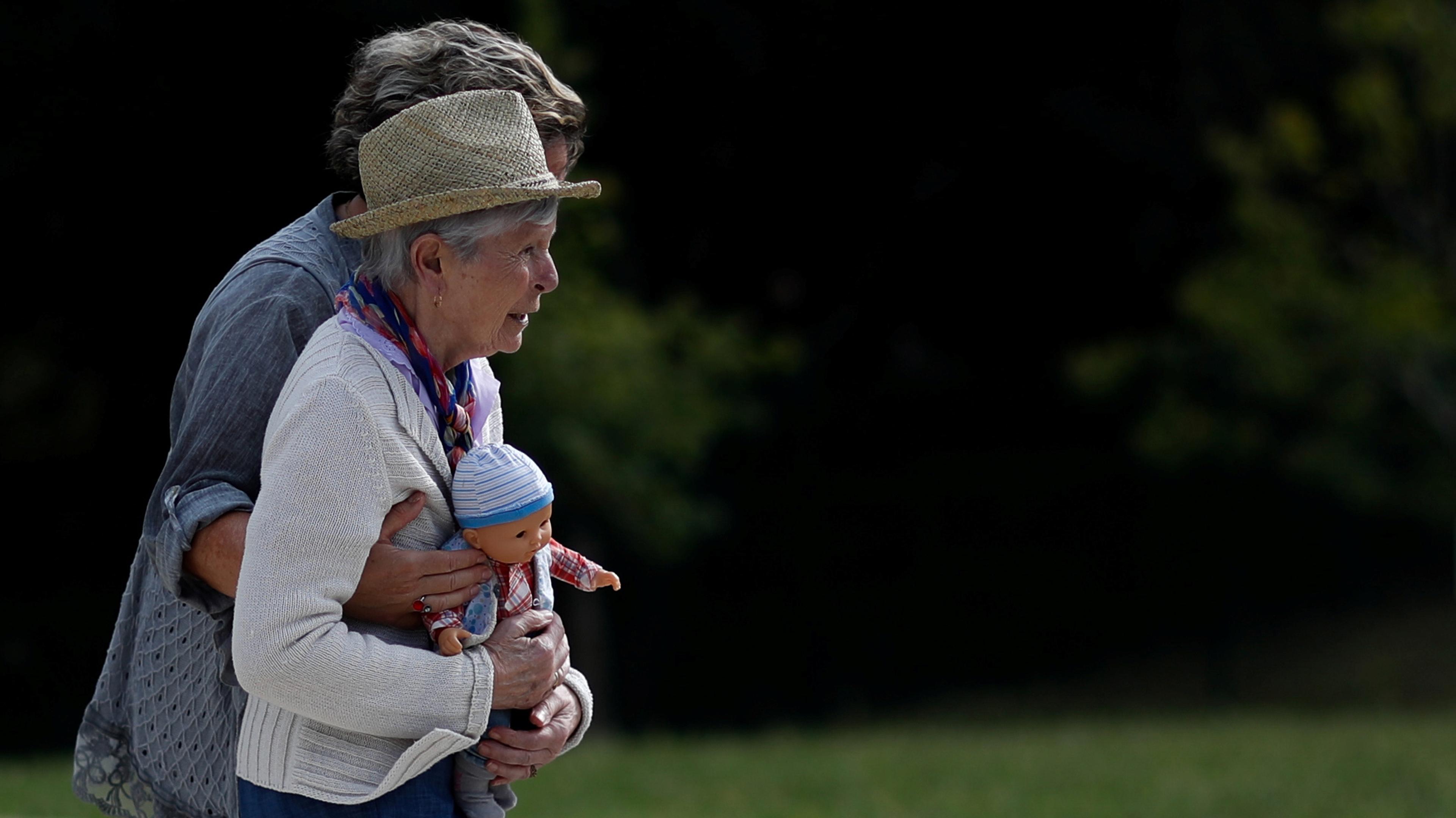 Photo of an older woman in a straw hat holding a doll in a park with another person embracing her from behind.