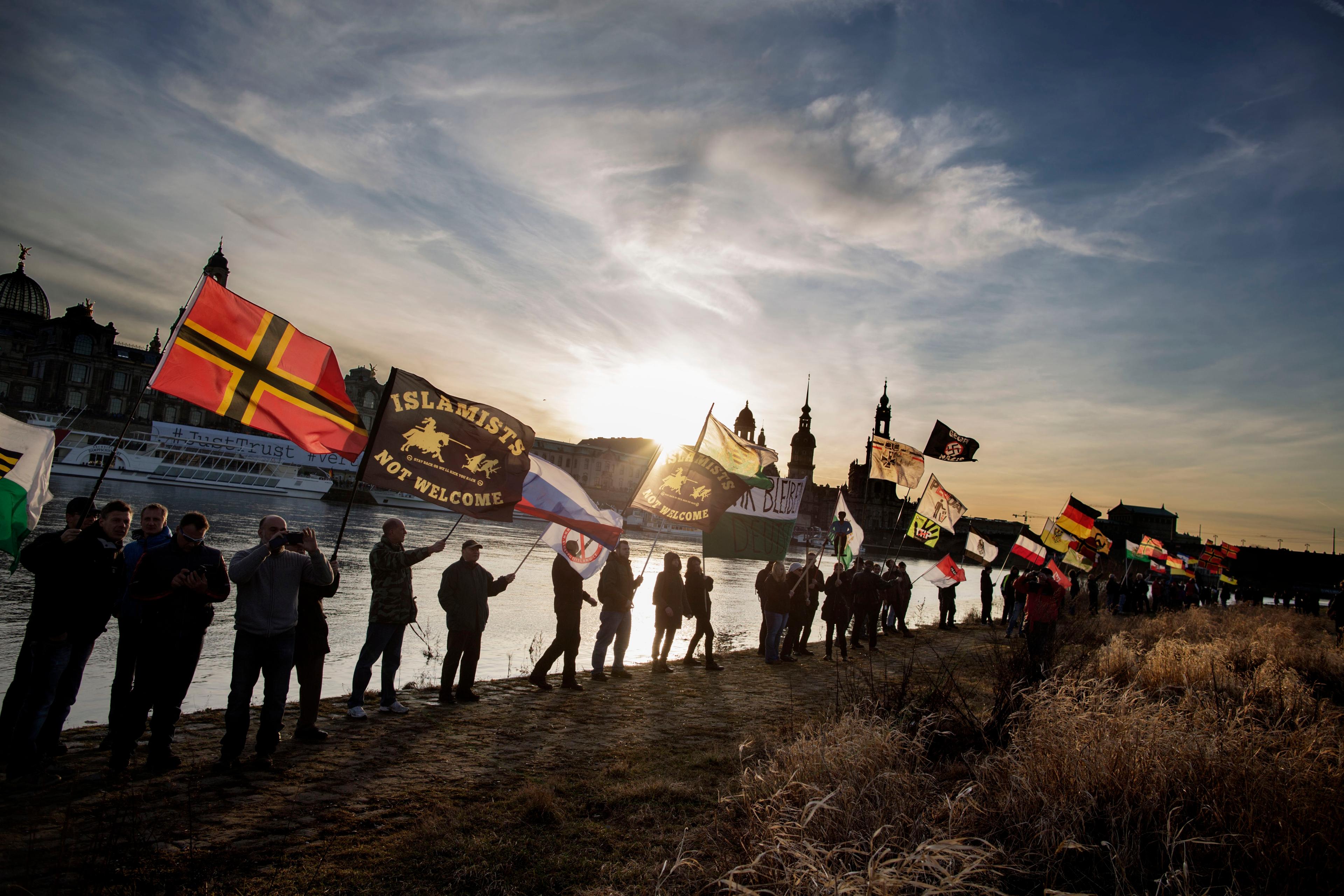 Photo of people holding flags with various messages along a riverside at sunset in an urban setting.