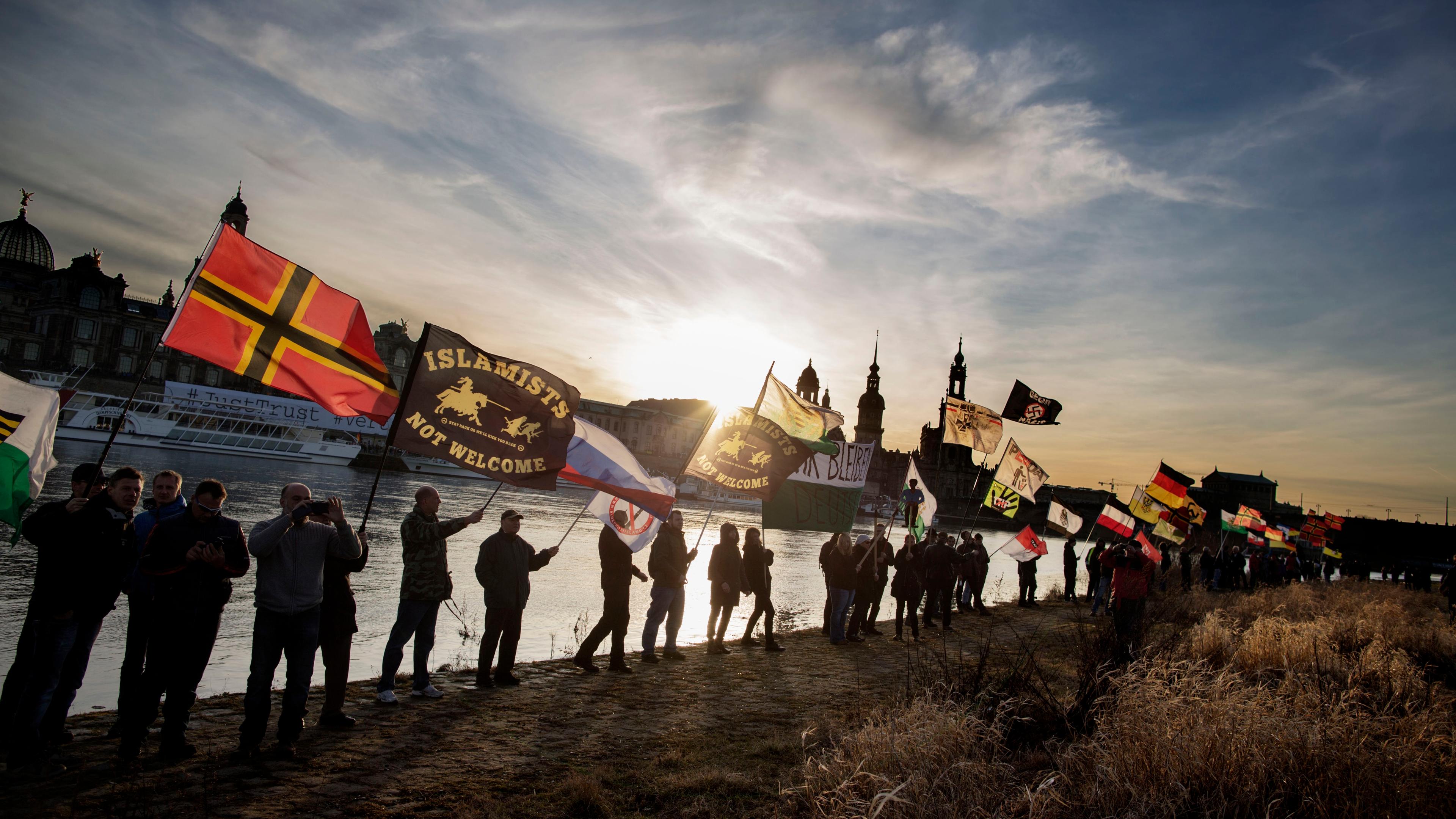 Photo of people holding flags with various messages along a riverside at sunset in an urban setting.