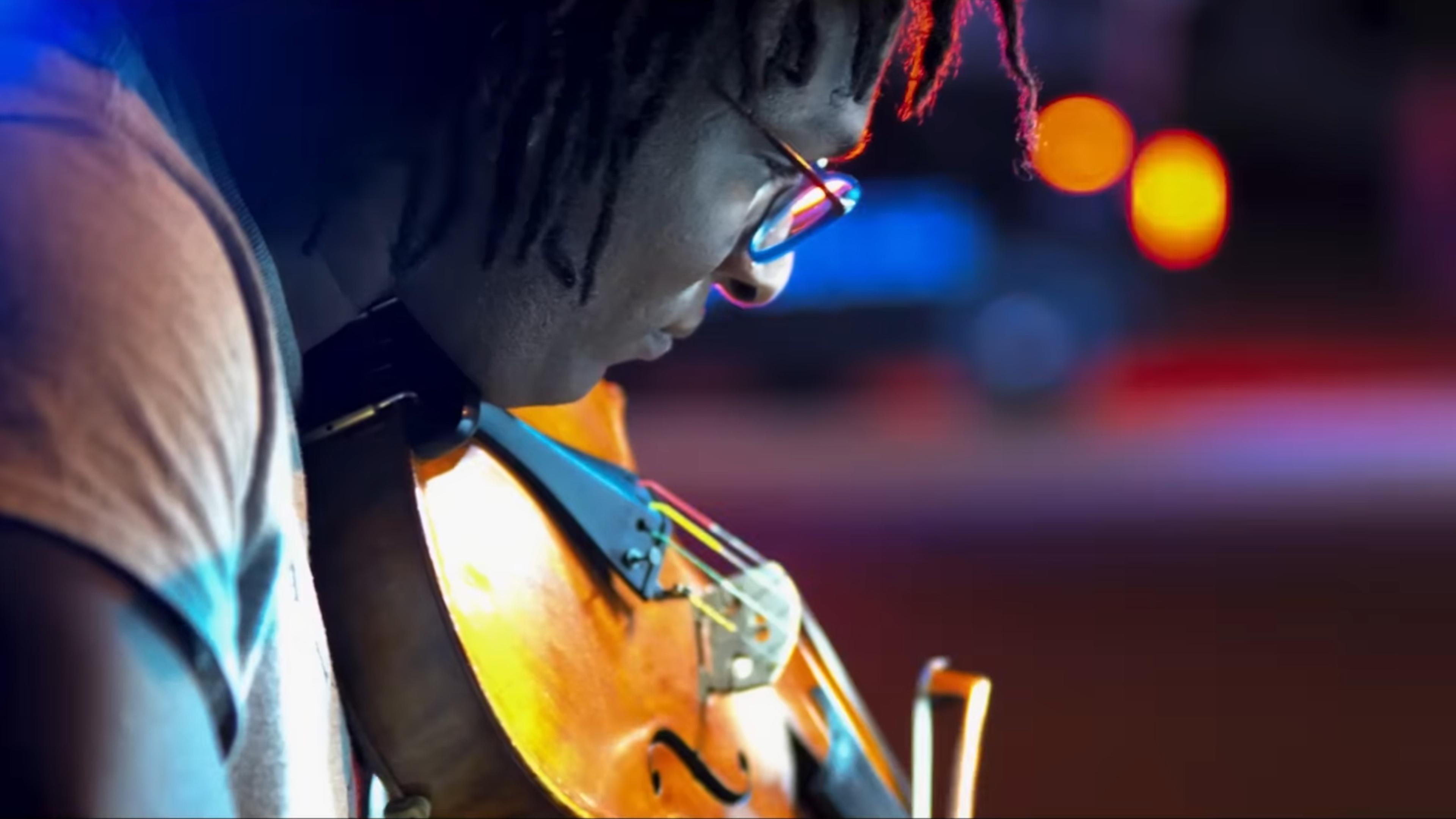 Close-up of a person with glasses playing the violin, illuminated by colourful lights in the background.