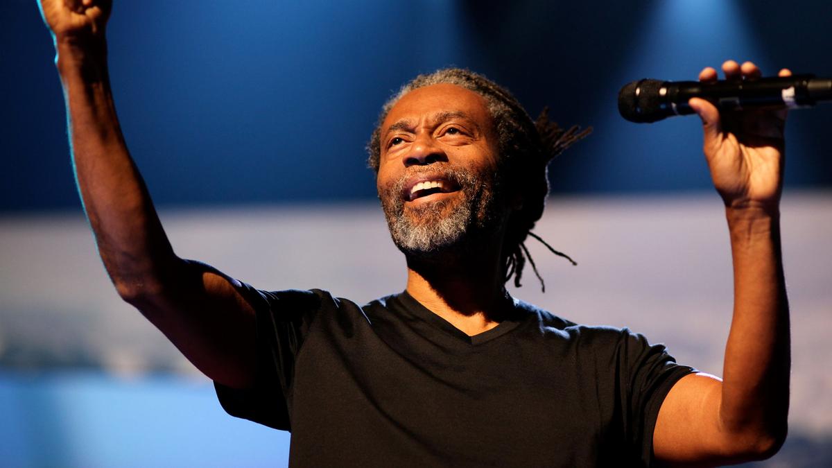A man with dreadlocks smiling, holding a microphone on stage with his other arm raised under blue stage lights.