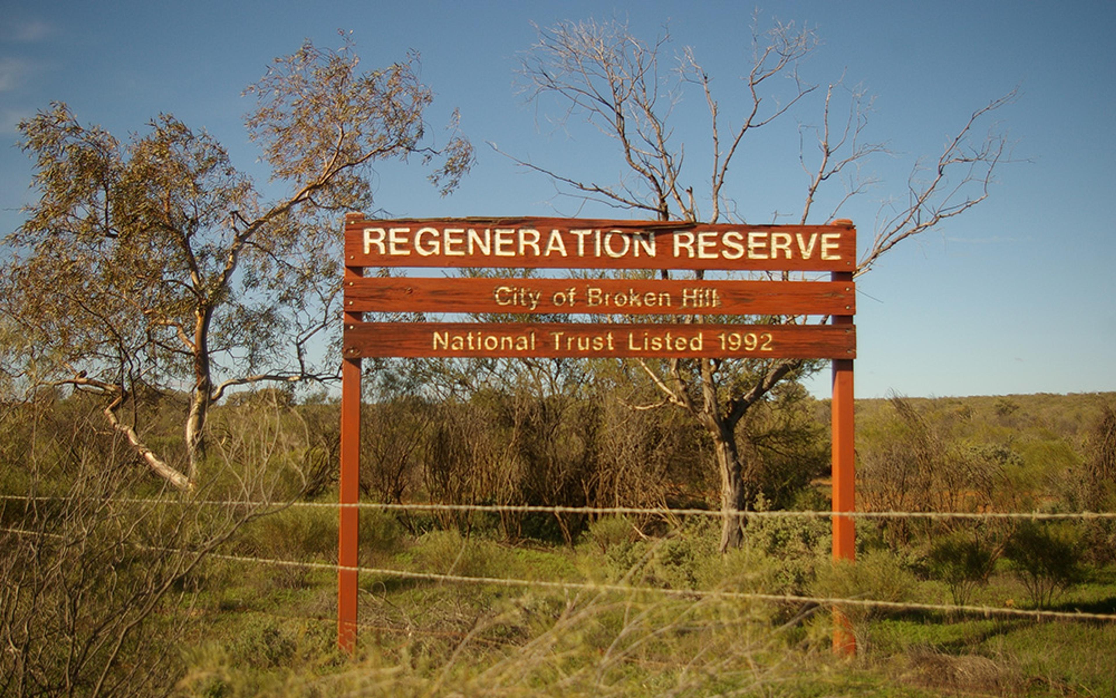 A sign reading ‘Regeneration Reserve City of Broken Hill National Trust Listed 1992’ with trees and shrubs in the background.