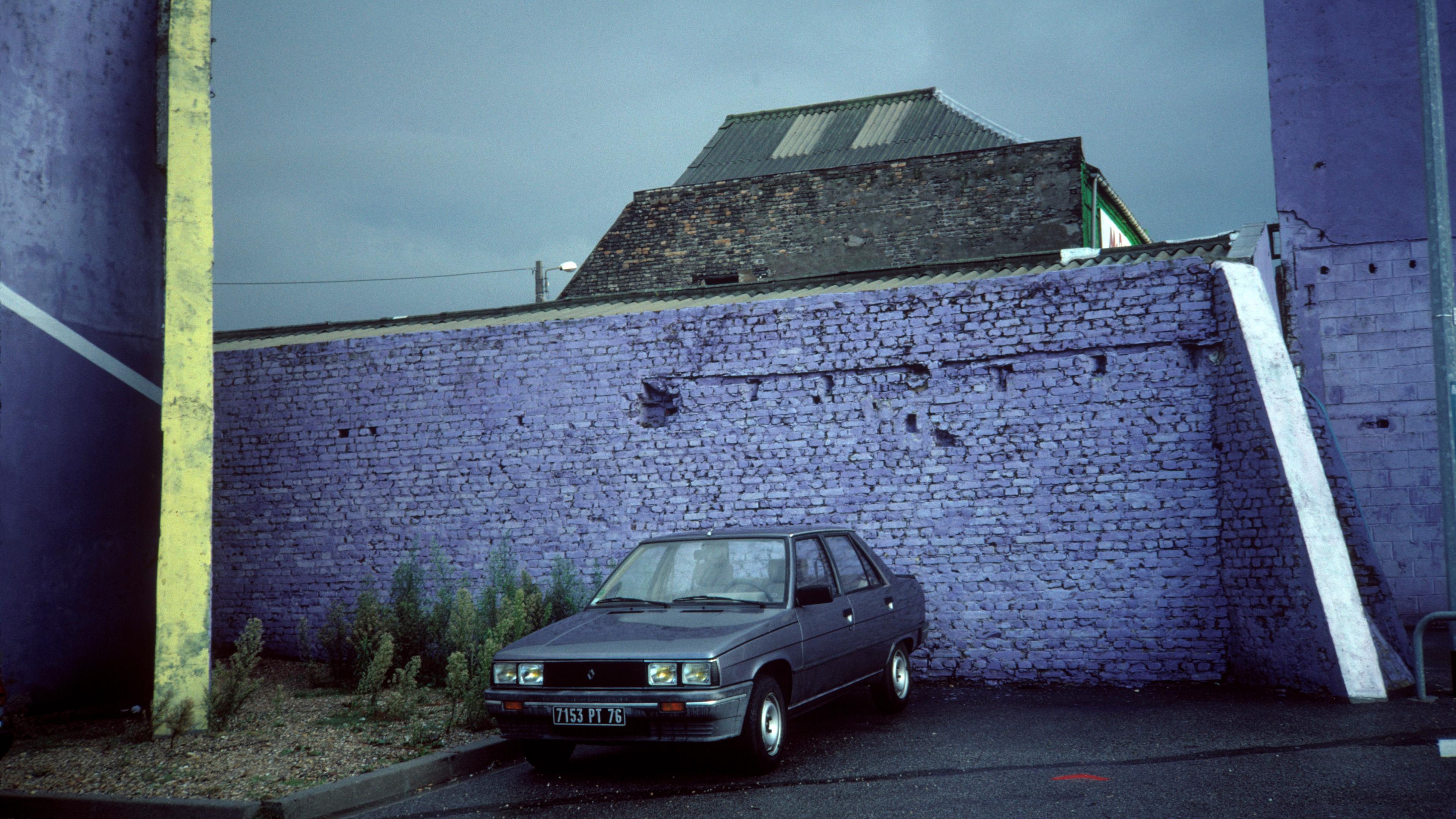 Photo of a grey car parked by a purple brick wall on a rainy day with an industrial building in the background.