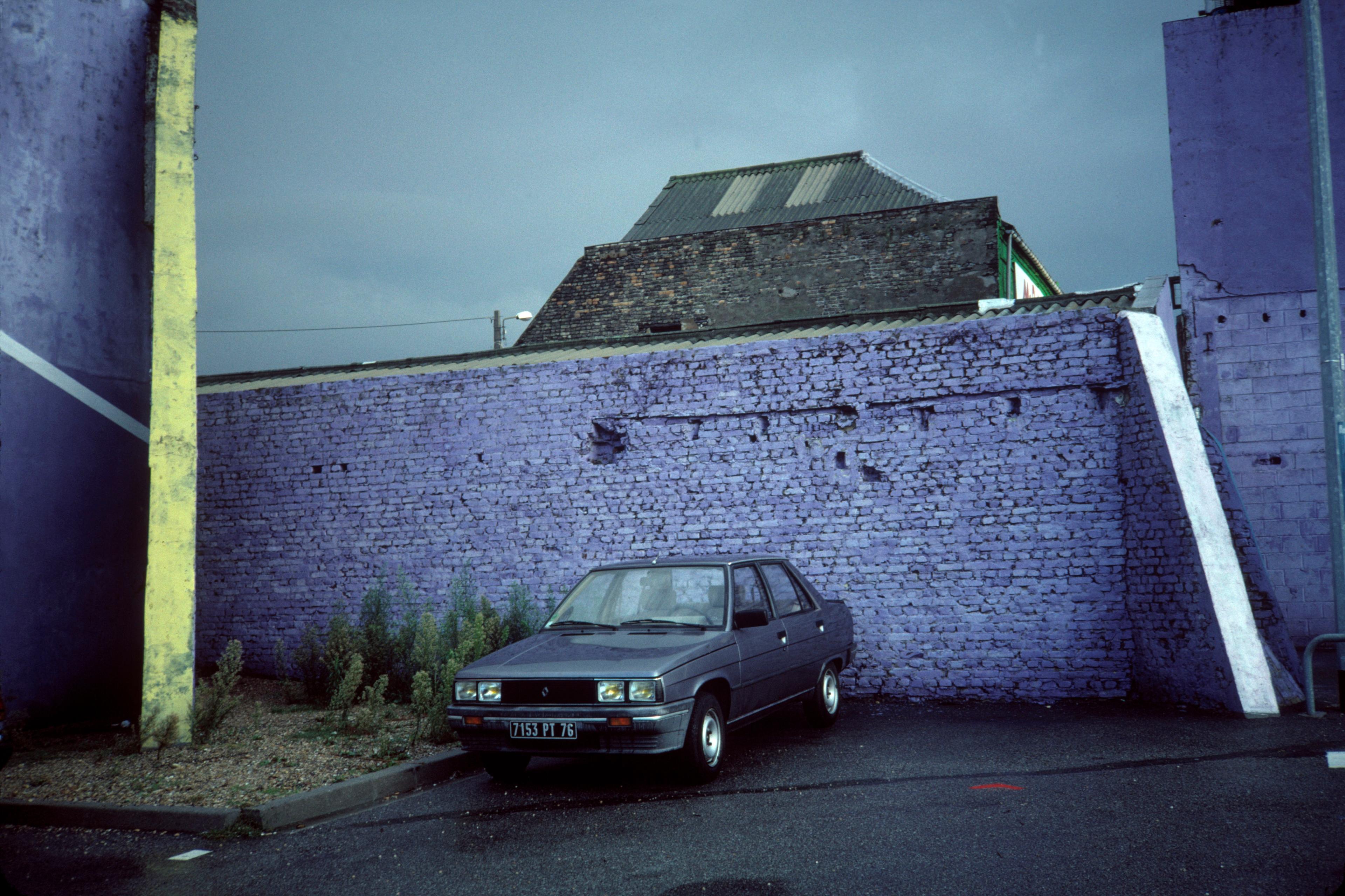 Photo of a grey car parked by a purple brick wall on a rainy day with an industrial building in the background.
