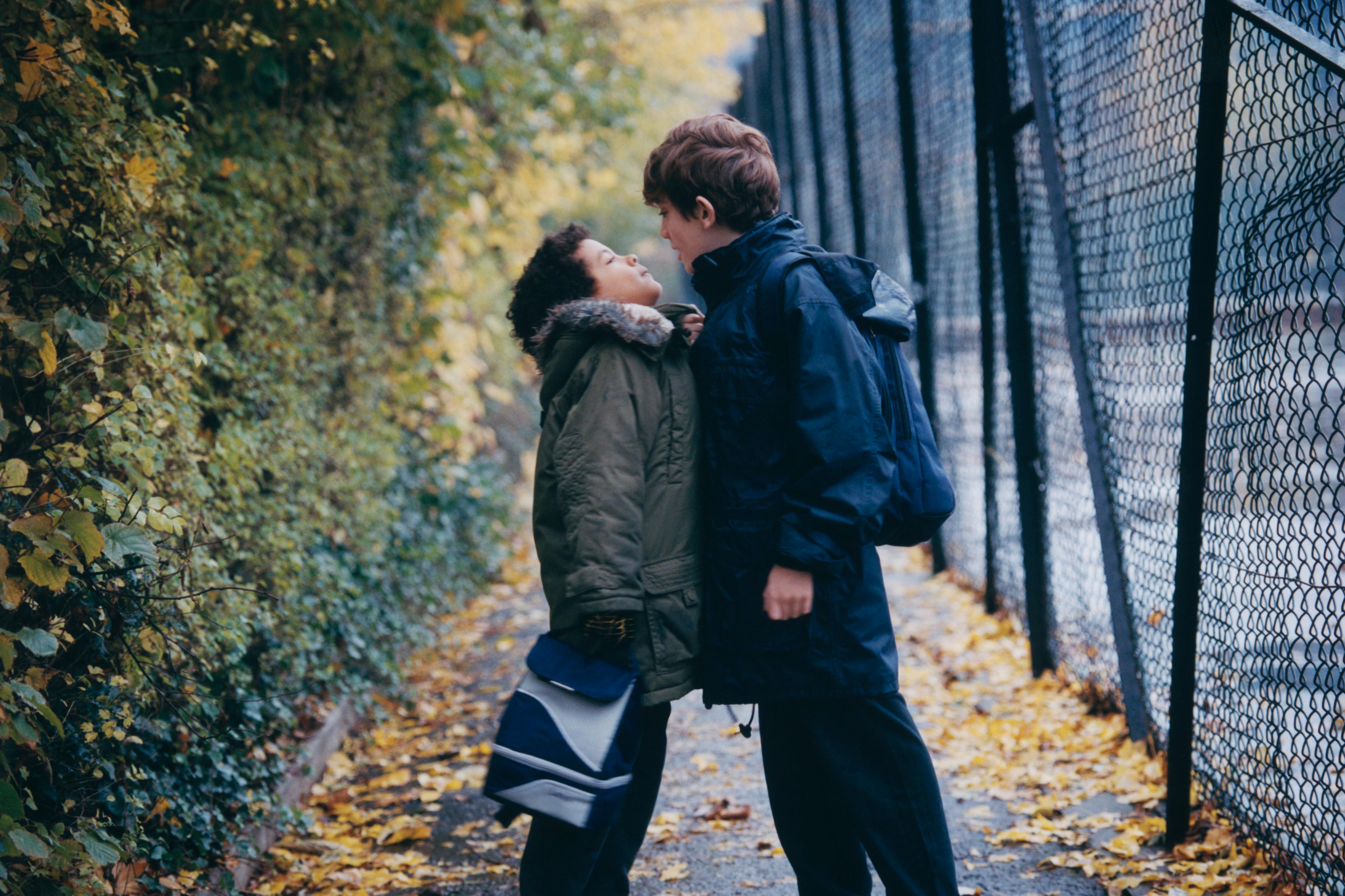 Photo of two boys playfully confronting each other on a leaf-covered path beside a fence in autumn.