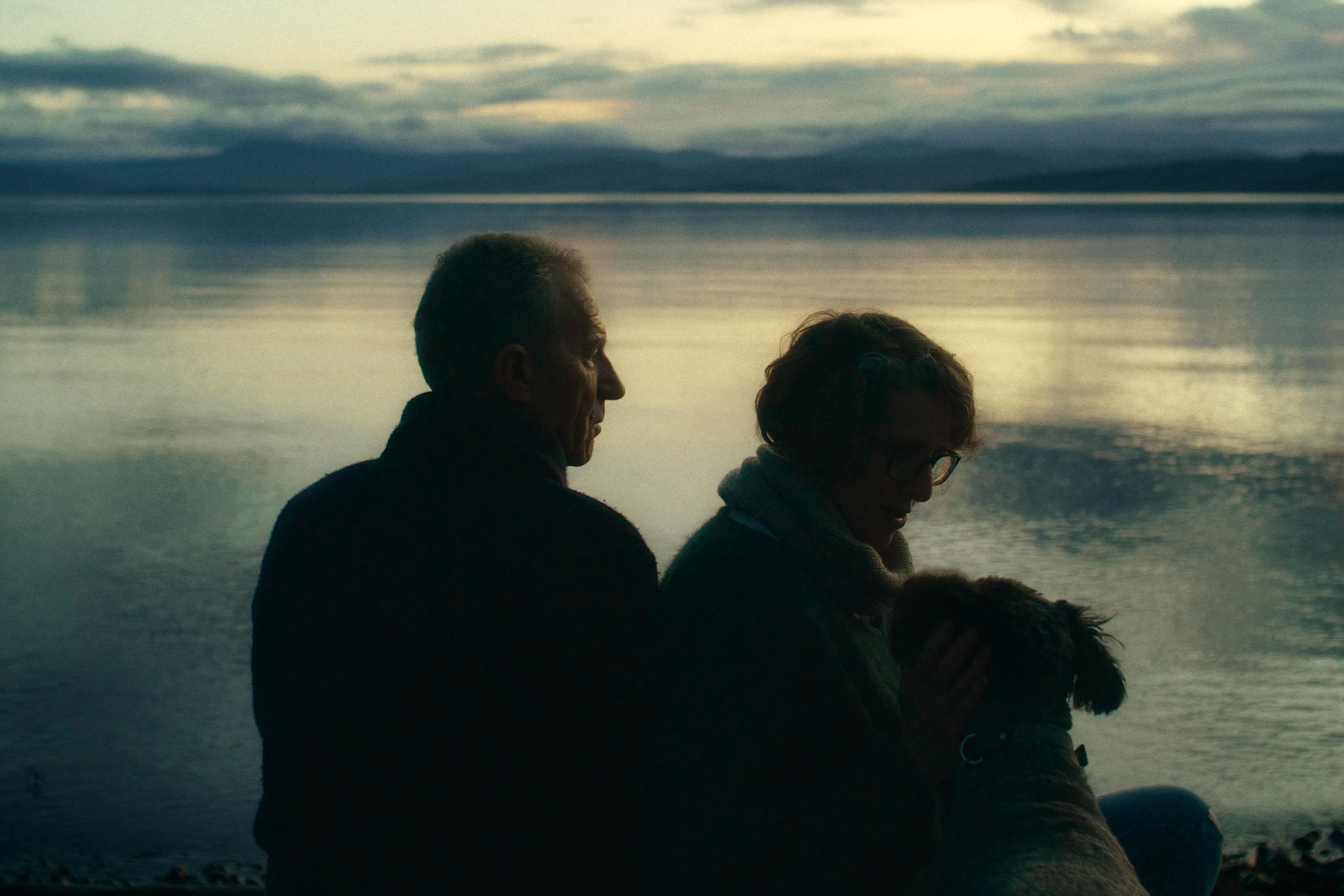 A man and woman sit by a calm lake during dusk, with mountains in the background and a dog resting near the woman.