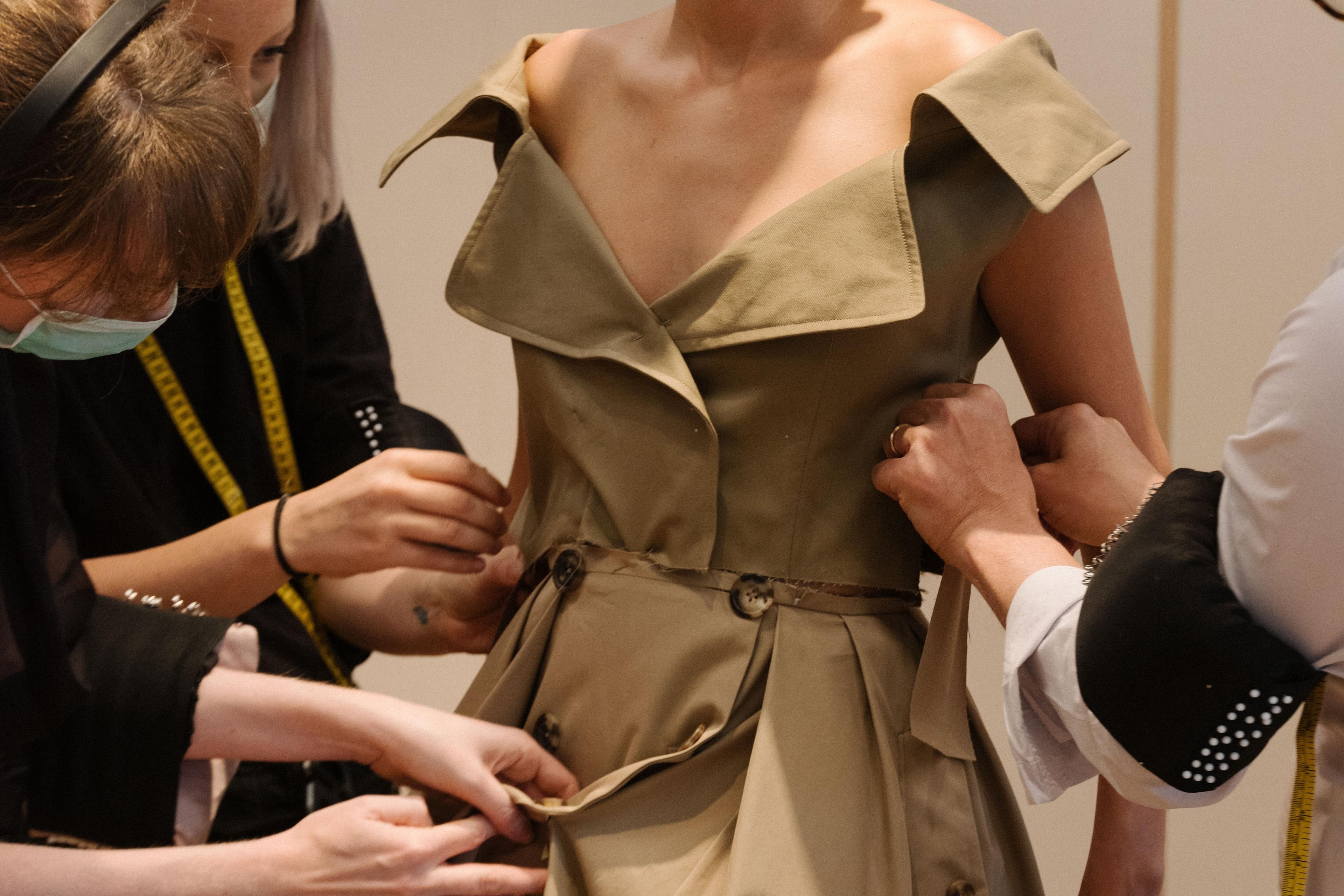 Photo of a woman wearing a tan dress being adjusted by three people, with focus on tailoring and fit adjustments.