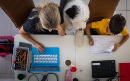 Woman working on a laptop, child drawing on paper, and a dog between them on a table with pencils and a coffee cup.