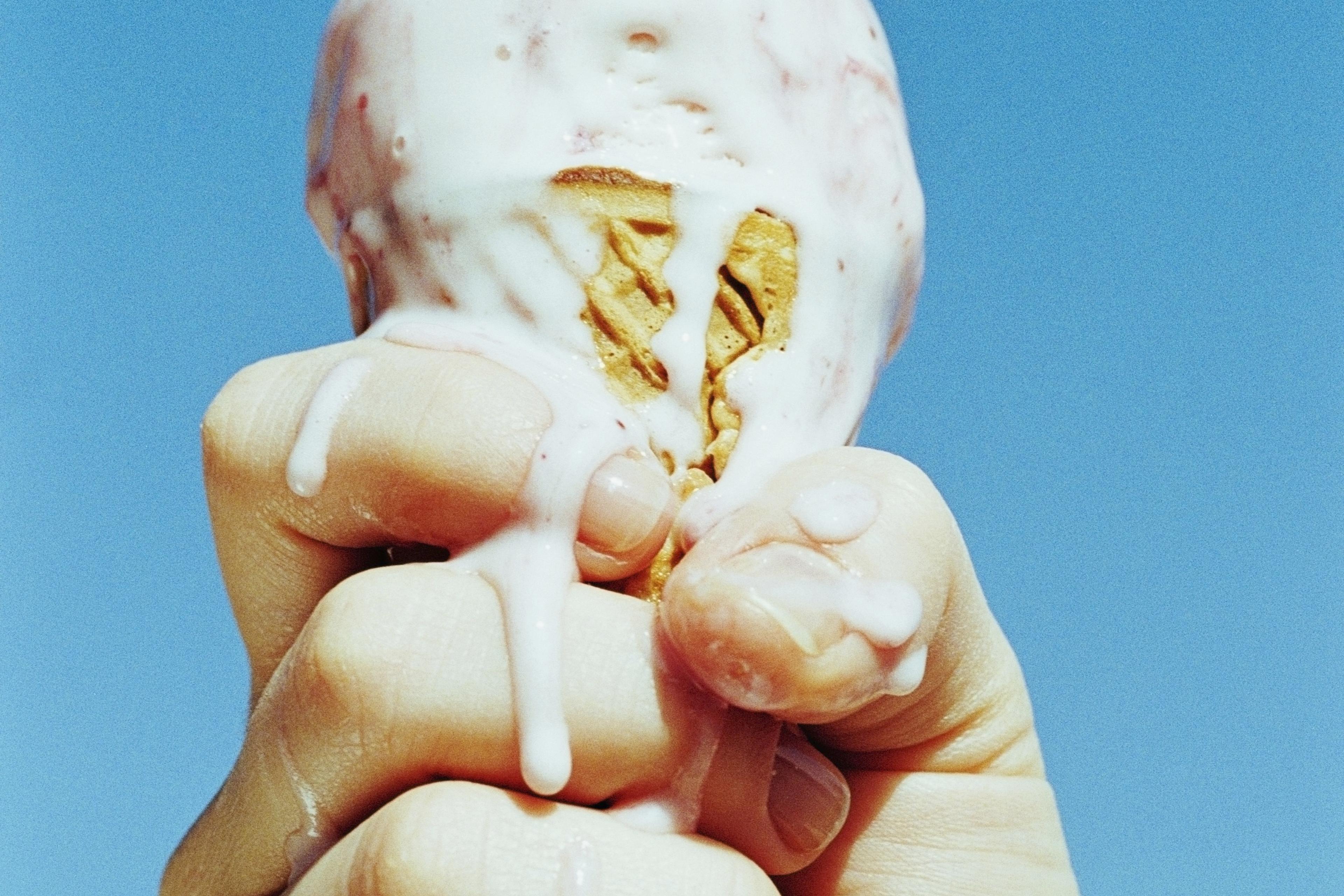 Photo of a hand holding a melting ice cream cone against a clear blue sky.