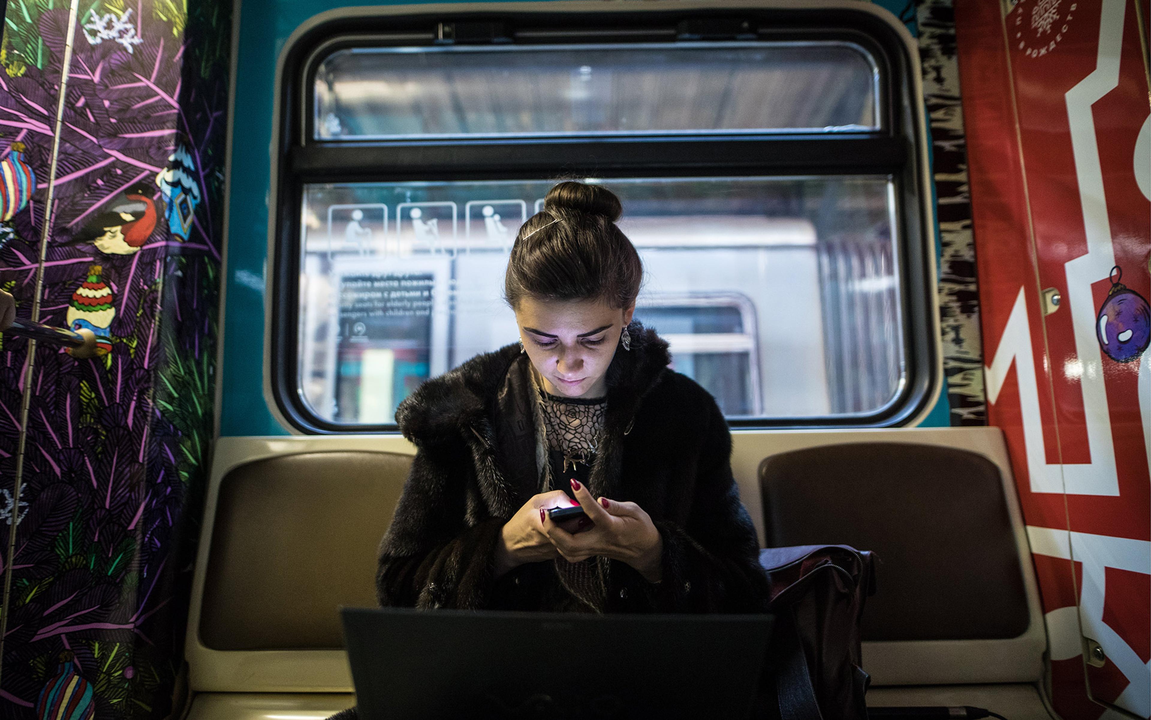 A woman using a smartphone on a train; colourful murals decorate the walls and an open laptop sits in her lap.