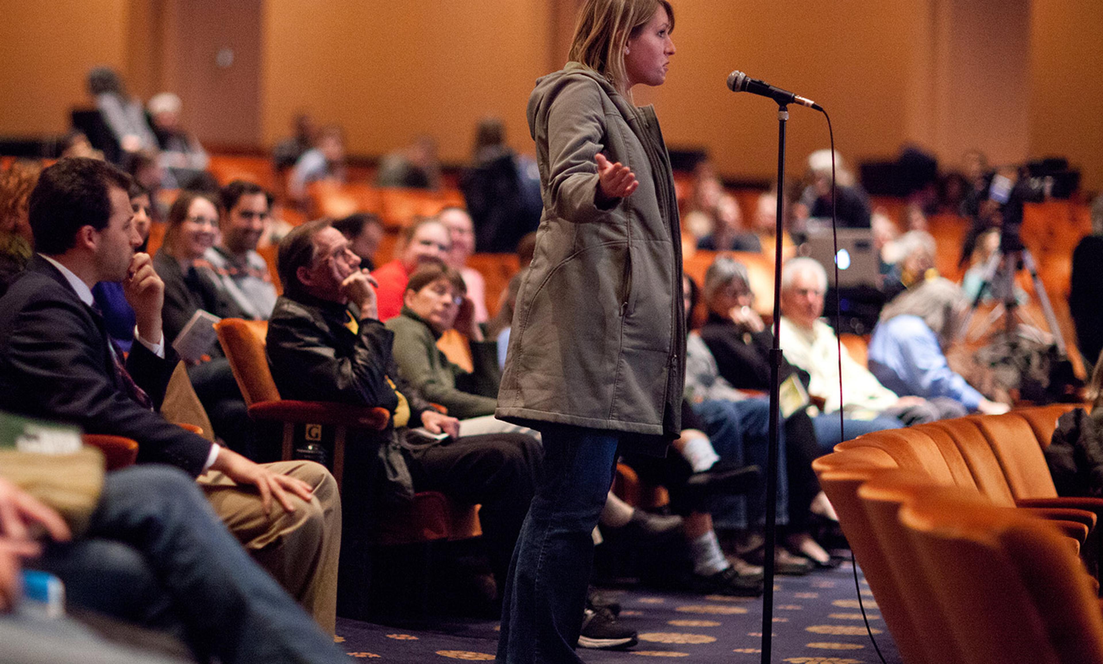 A woman speaking into a microphone from the audience in an auditorium. People are seated and listening attentively.