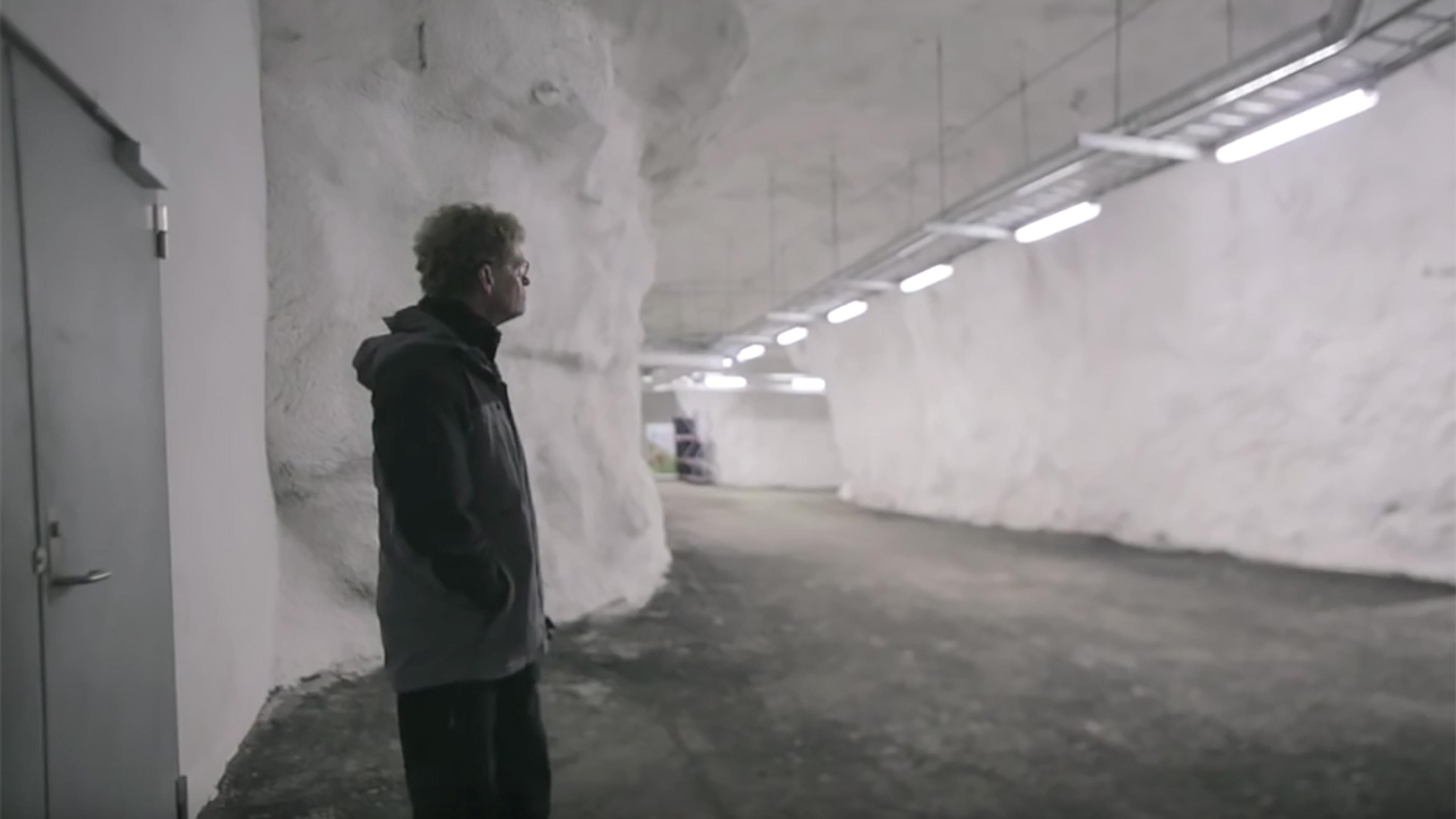 A man in a large, well-lit, underground tunnel with white walls and industrial ceiling lights, facing away towards the tunnel.