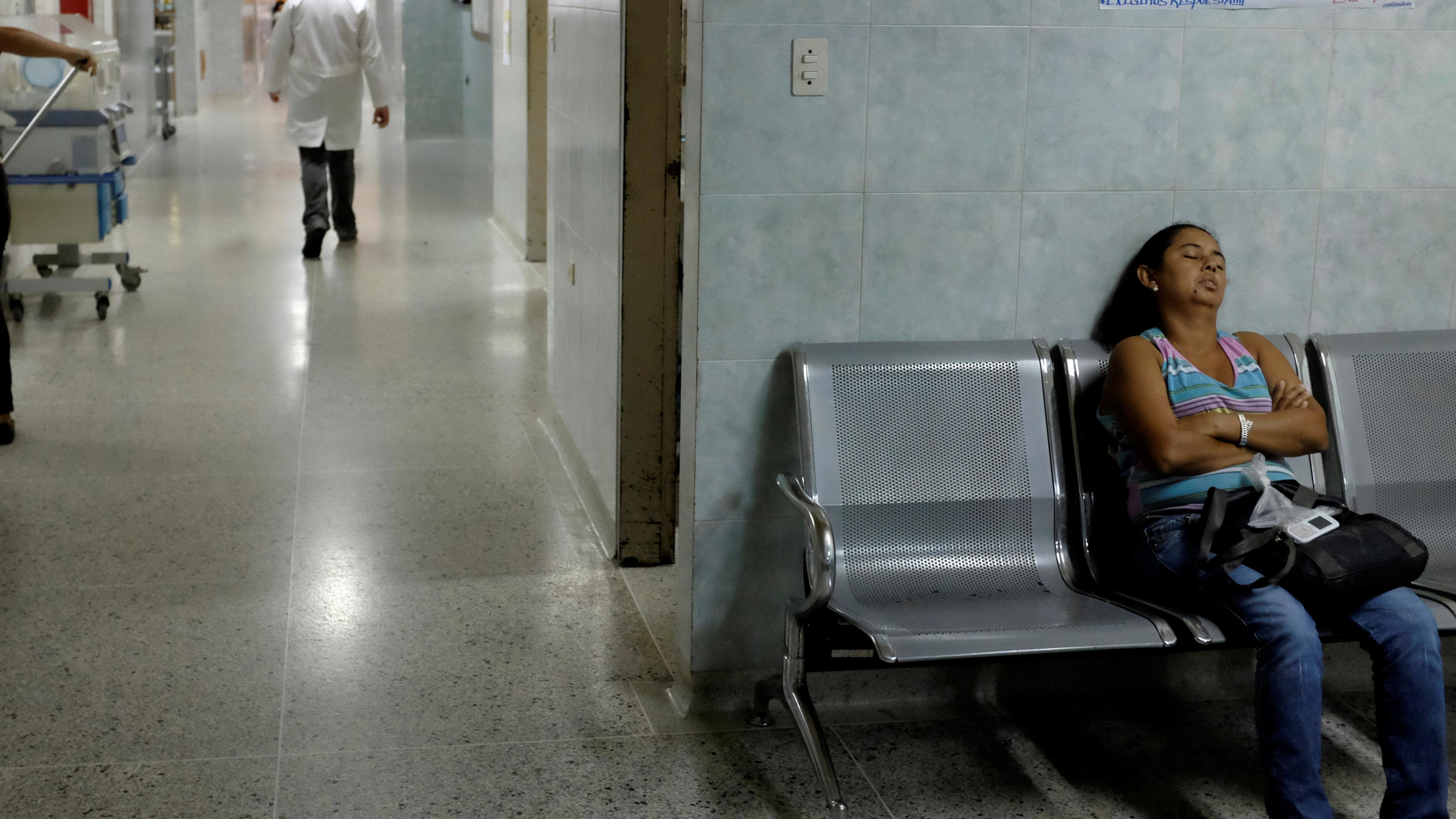 A hospital corridor with a woman asleep on a bench and a doctor walking away in the background.