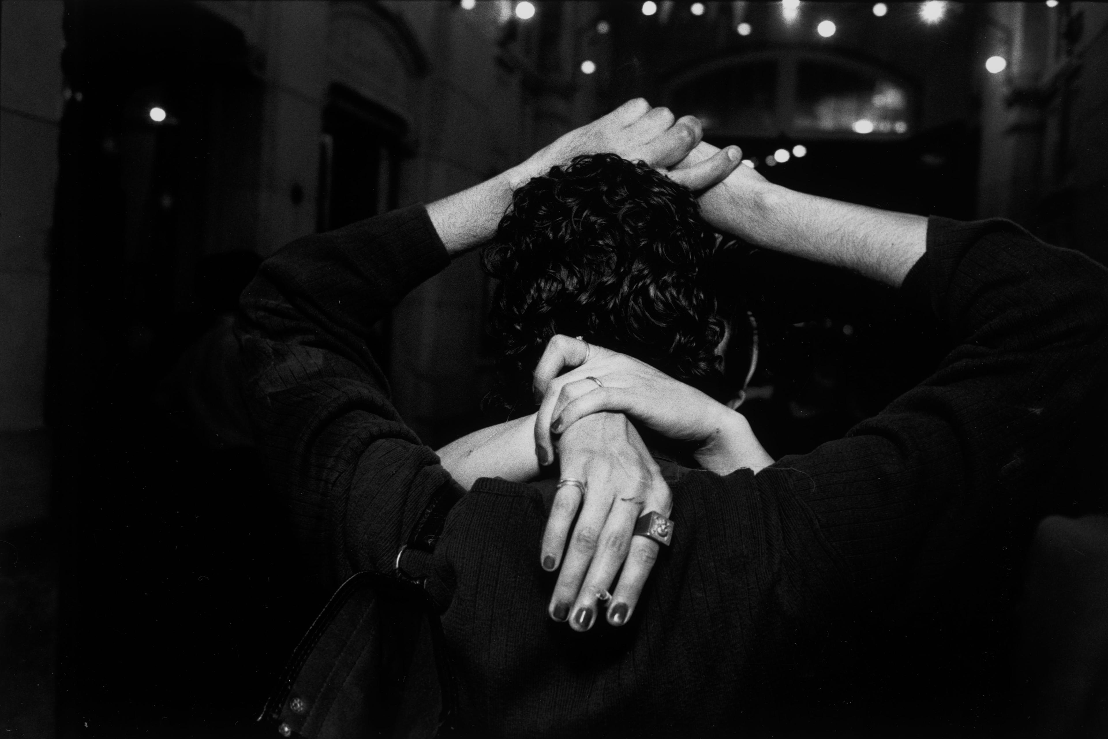 Black and white photo of a couple embracing with their hands intertwined behind one person’s head, set in a dimly lit space.