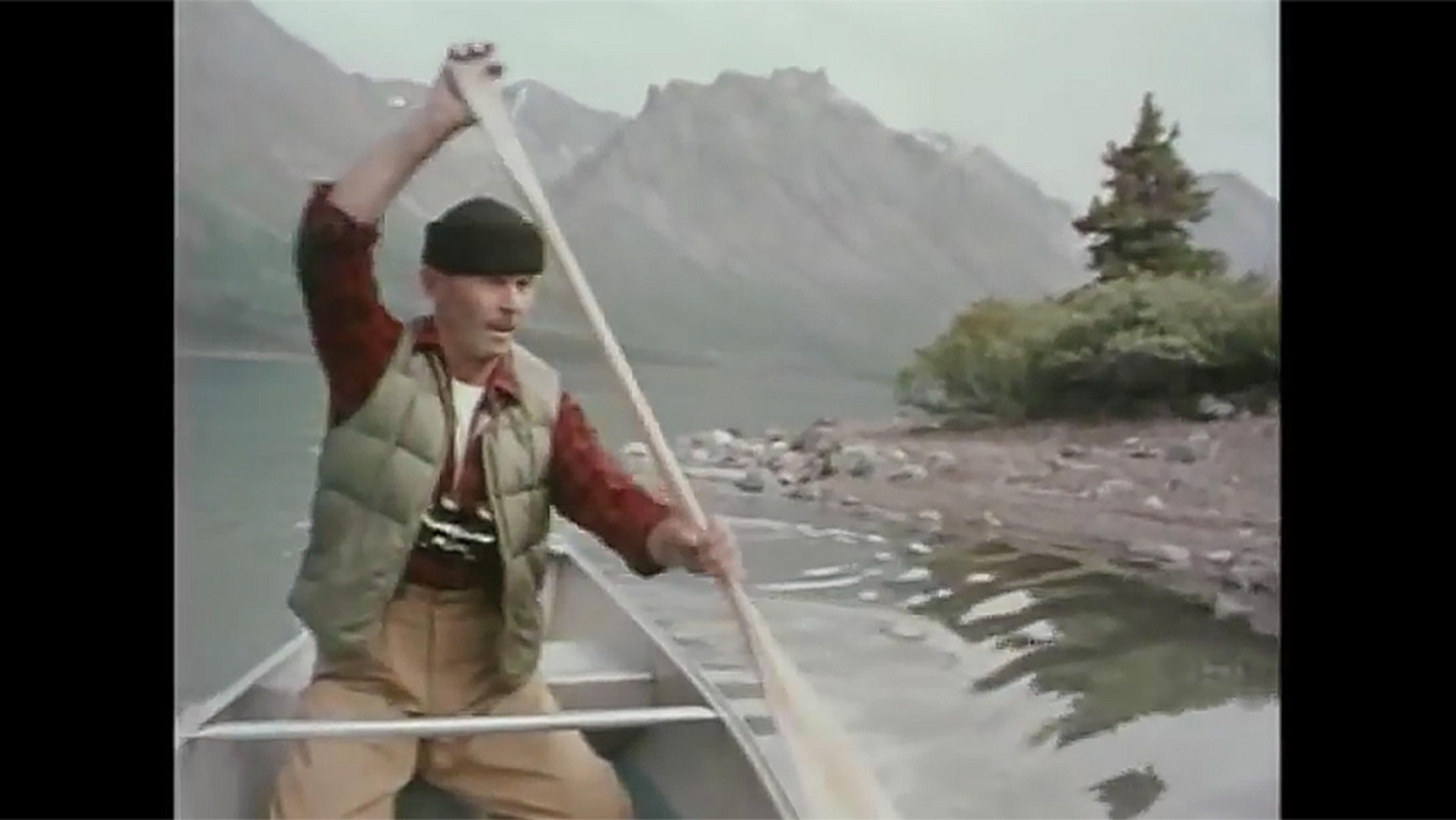 A man in a beanie and vest paddling a canoe on a calm lake with mountains and a tree in the background.