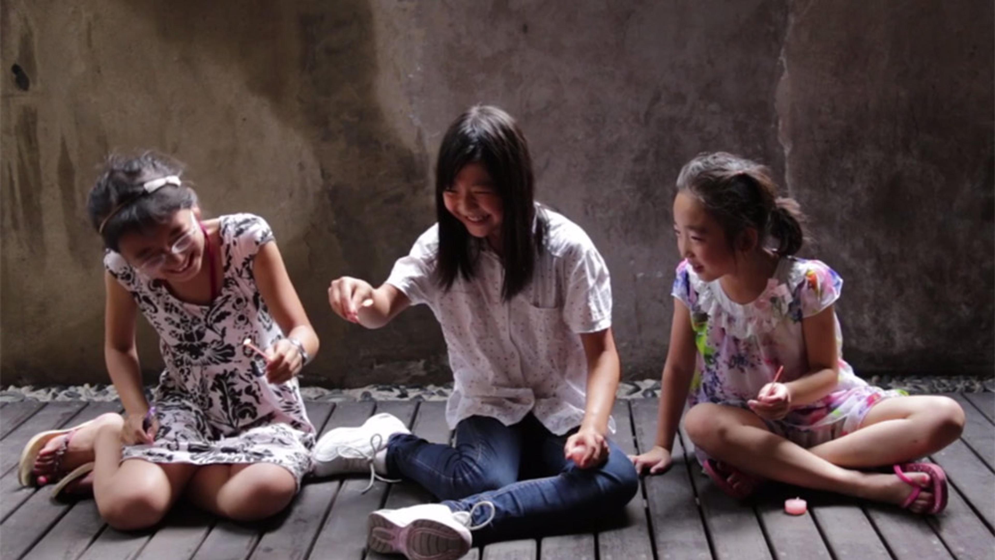 Three young Asian girls sitting on a wooden floor laughing and playing with small objects indoors against a concrete wall.