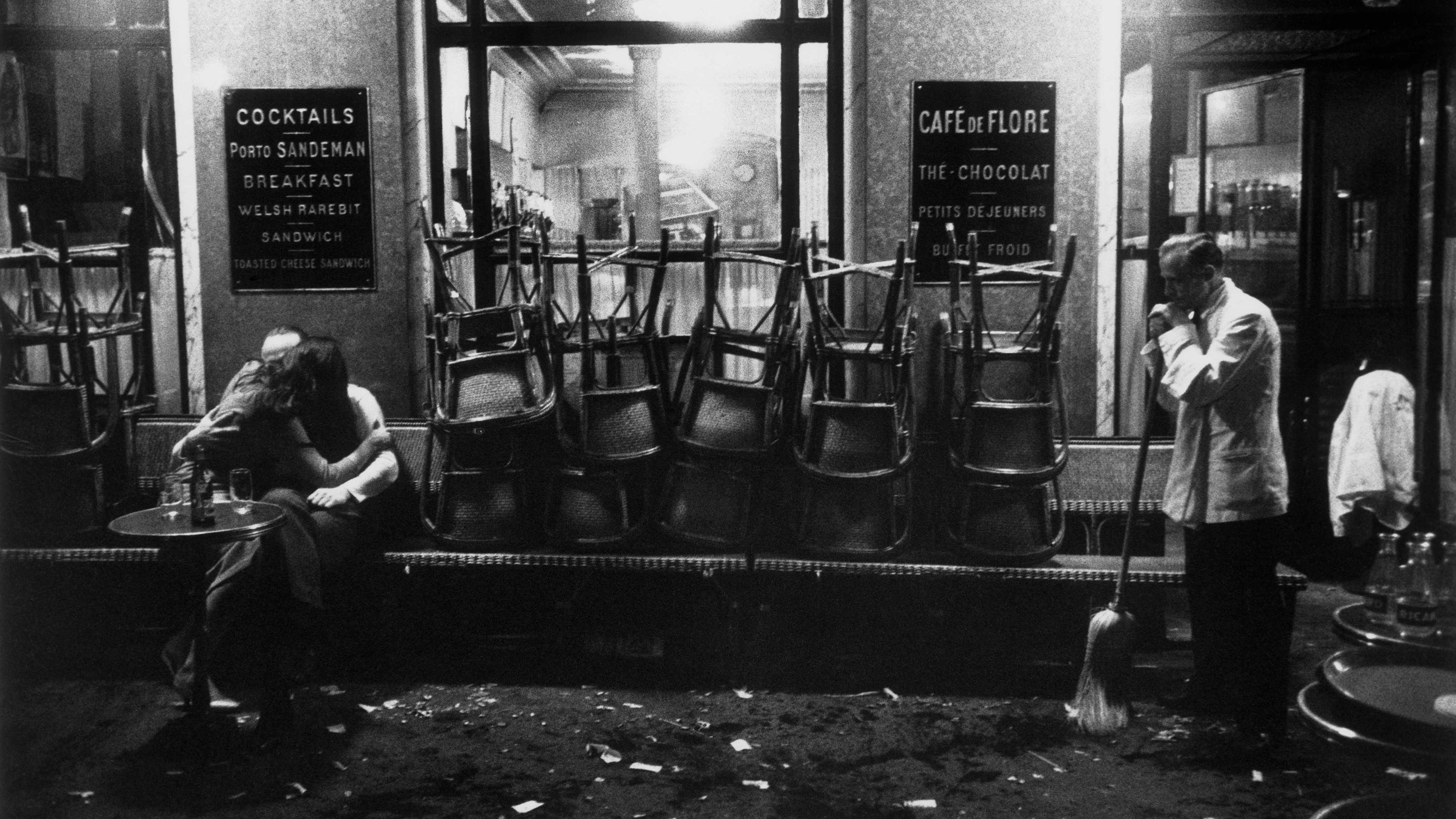 Black and white photo of a couple kissing outside a café at night with a waiter sweeping the floor.