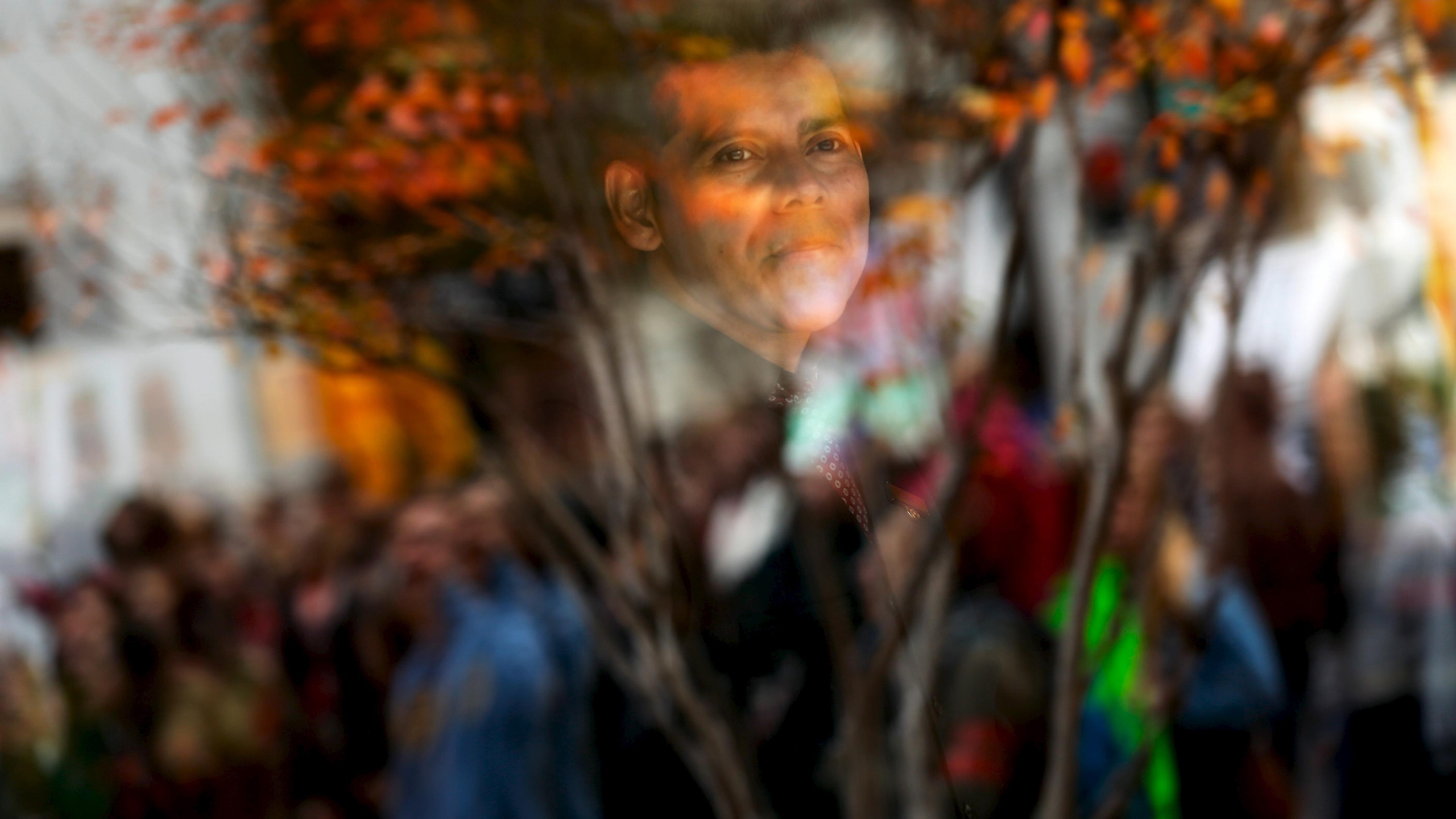 Photo showing a man’s reflection in glass with blurred people and tree branches in the foreground.