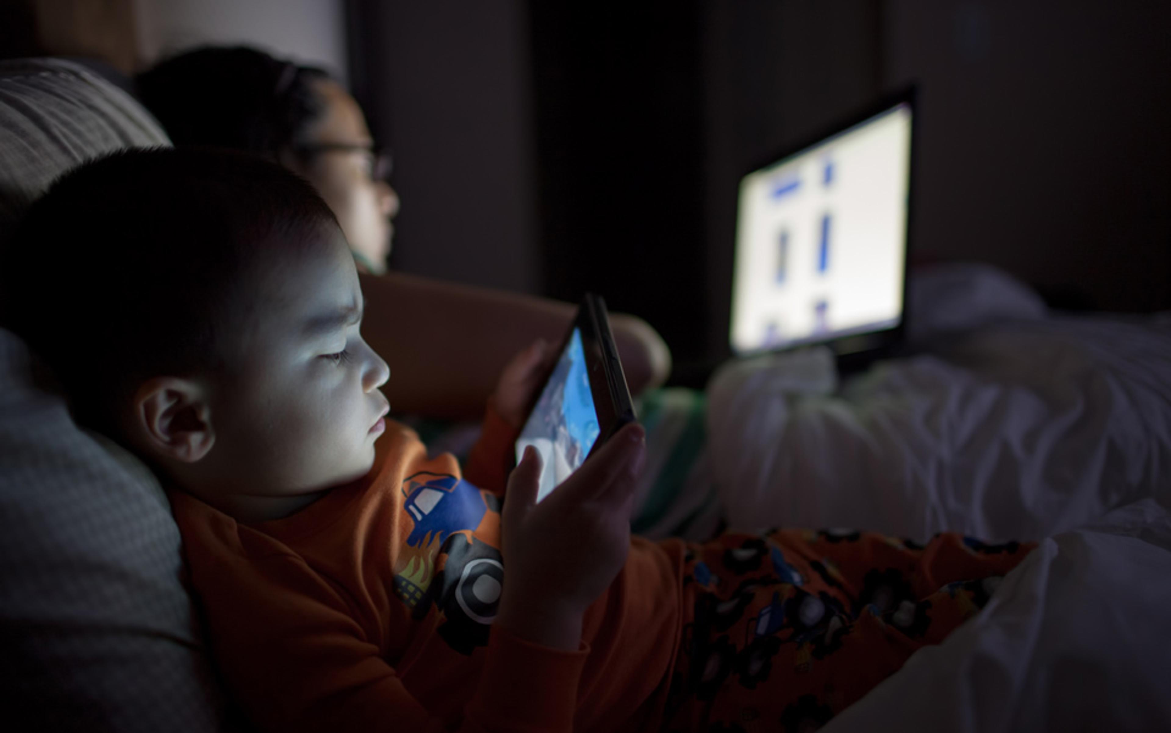 A young child in orange pyjamas using a tablet on a bed at night with an adult in the background using a laptop.