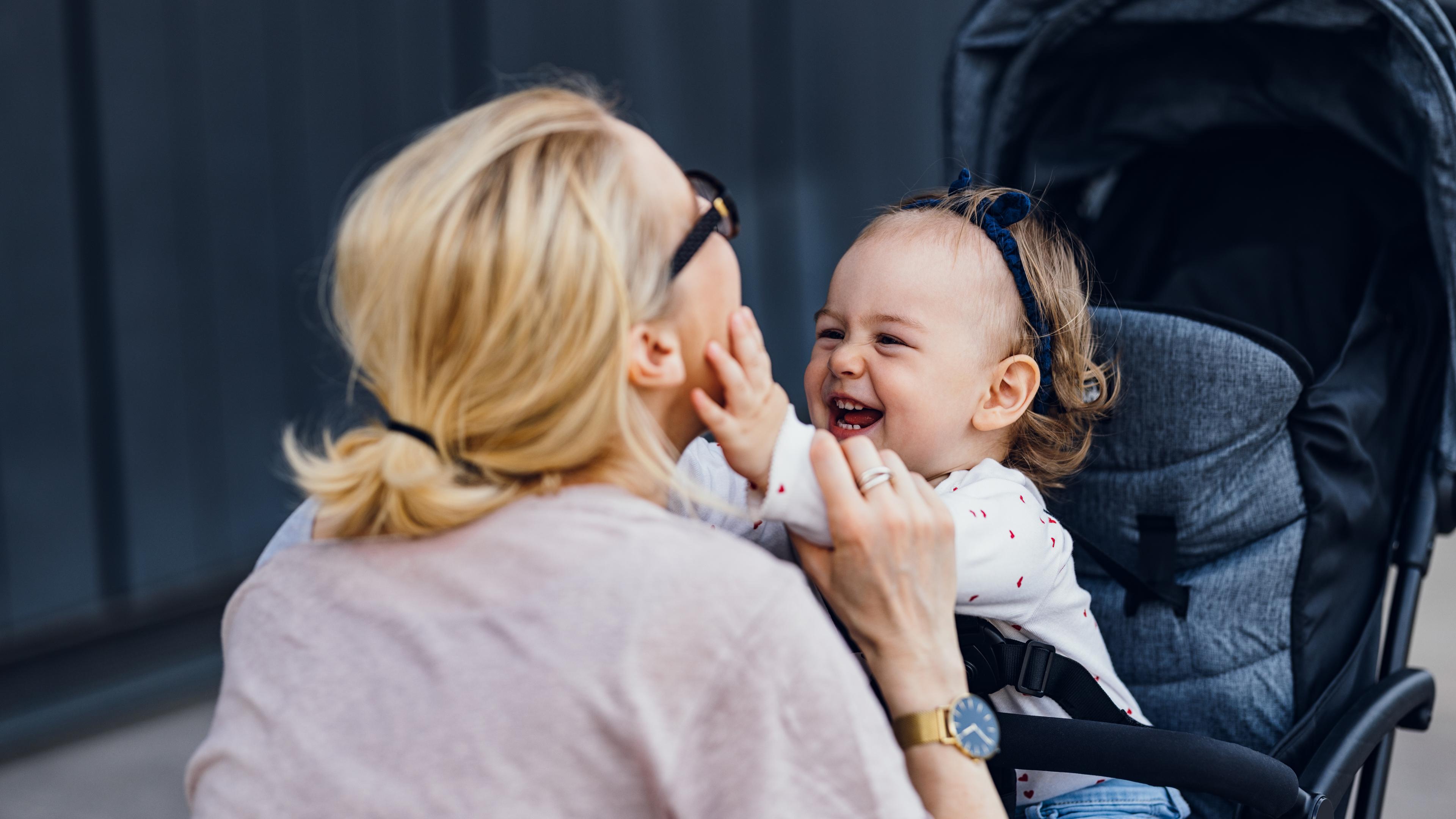 Photo of a woman playing with a smiling baby in a pram outside. The woman wears glasses and has her hair tied back.