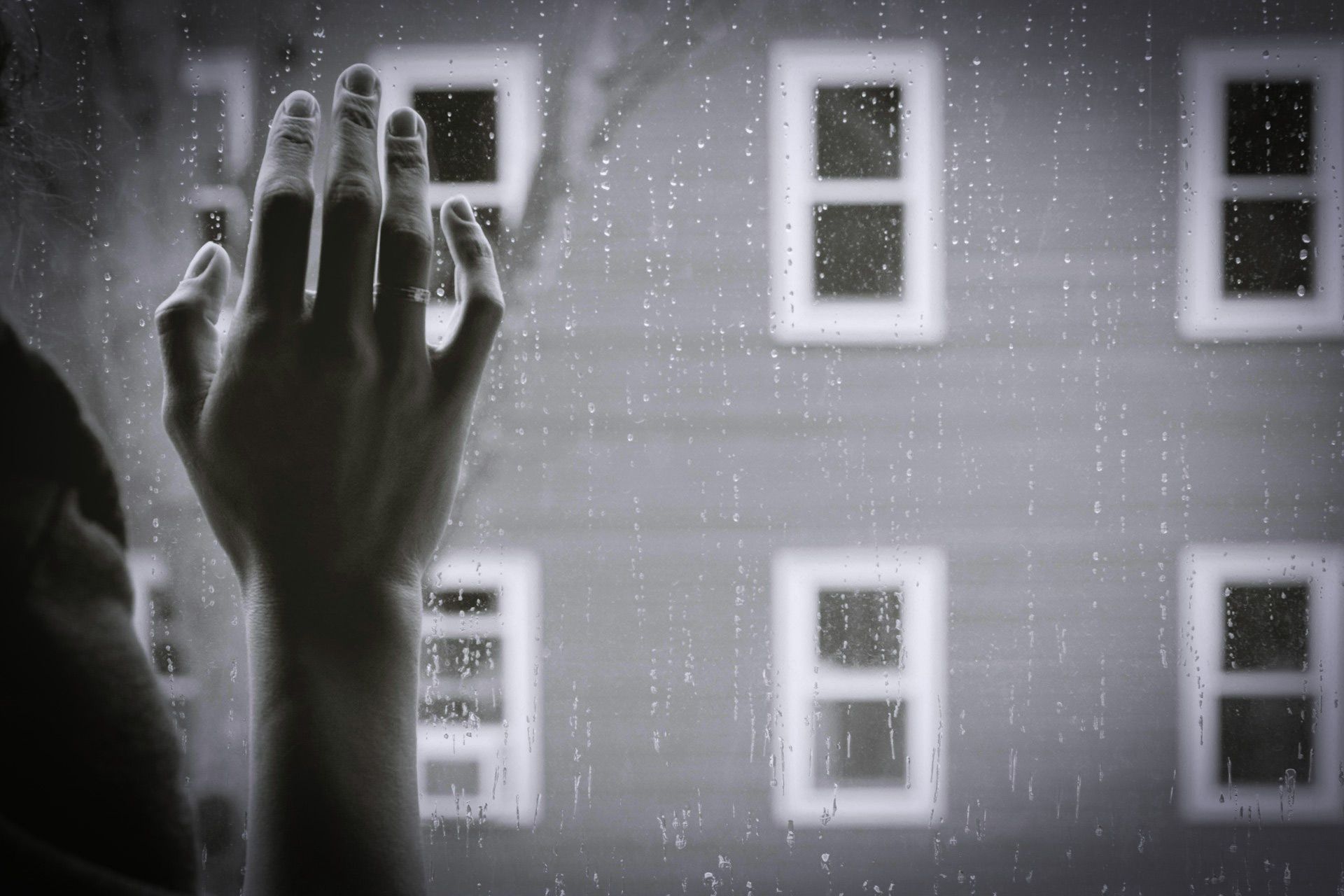 Black and white photo of a hand touching a rain-specked window with four blurred windows in the background.