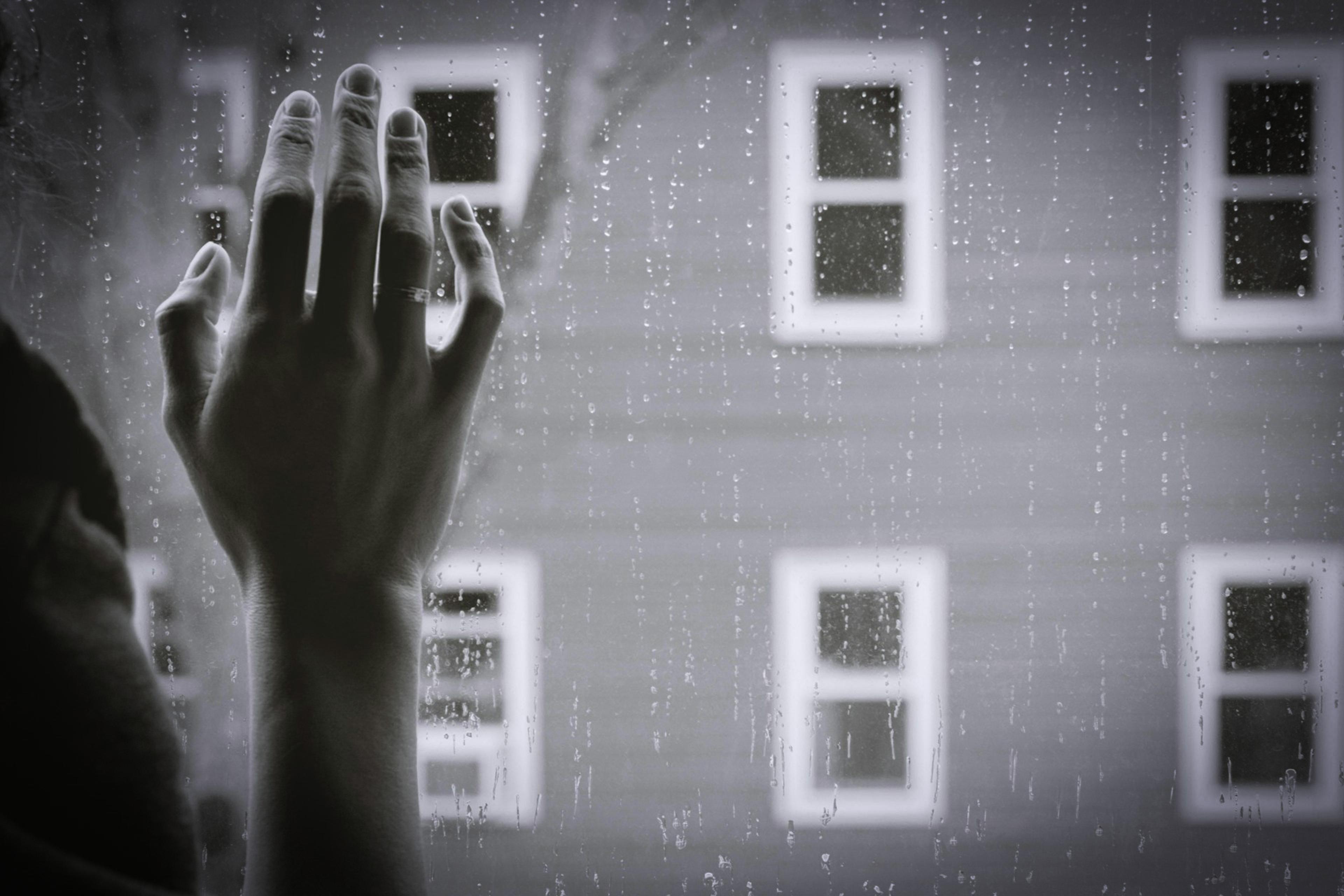 Black and white photo of a hand touching a rain-specked window with four blurred windows in the background.
