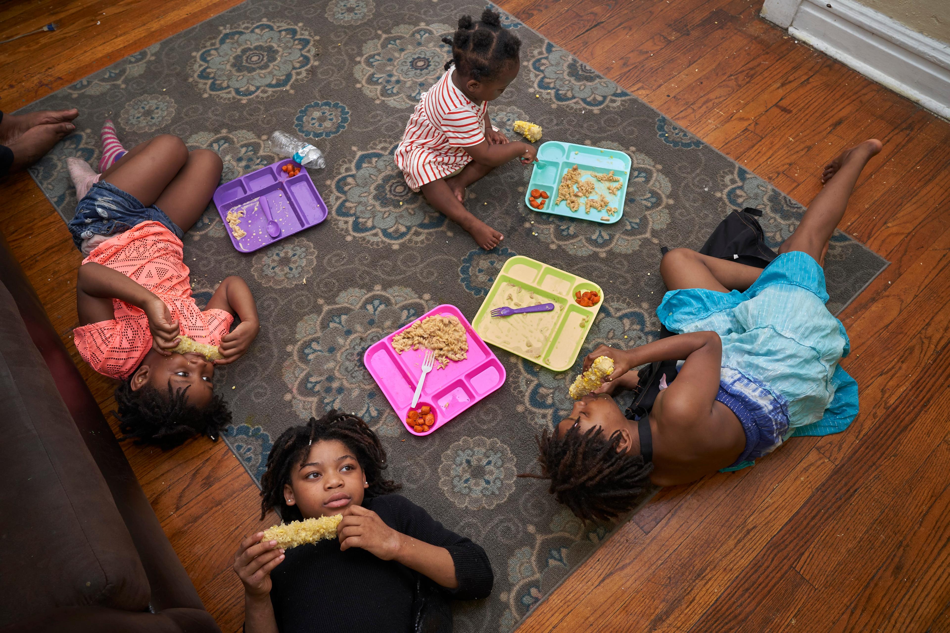 Photo of four children lying on a rug eating from colourful trays on a wooden floor with a patterned carpet.