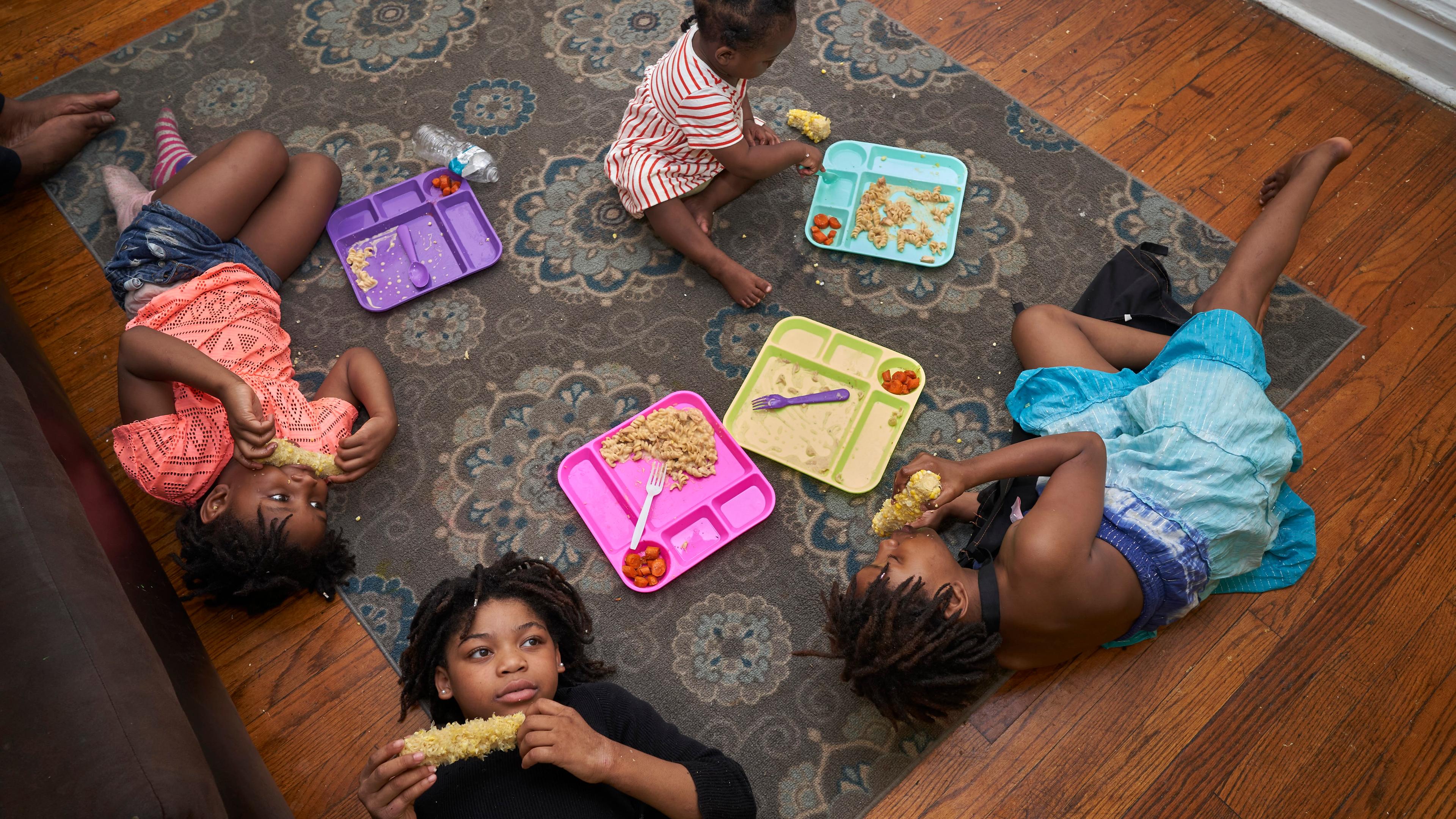 Photo of four children lying on a rug eating from colourful trays on a wooden floor with a patterned carpet.