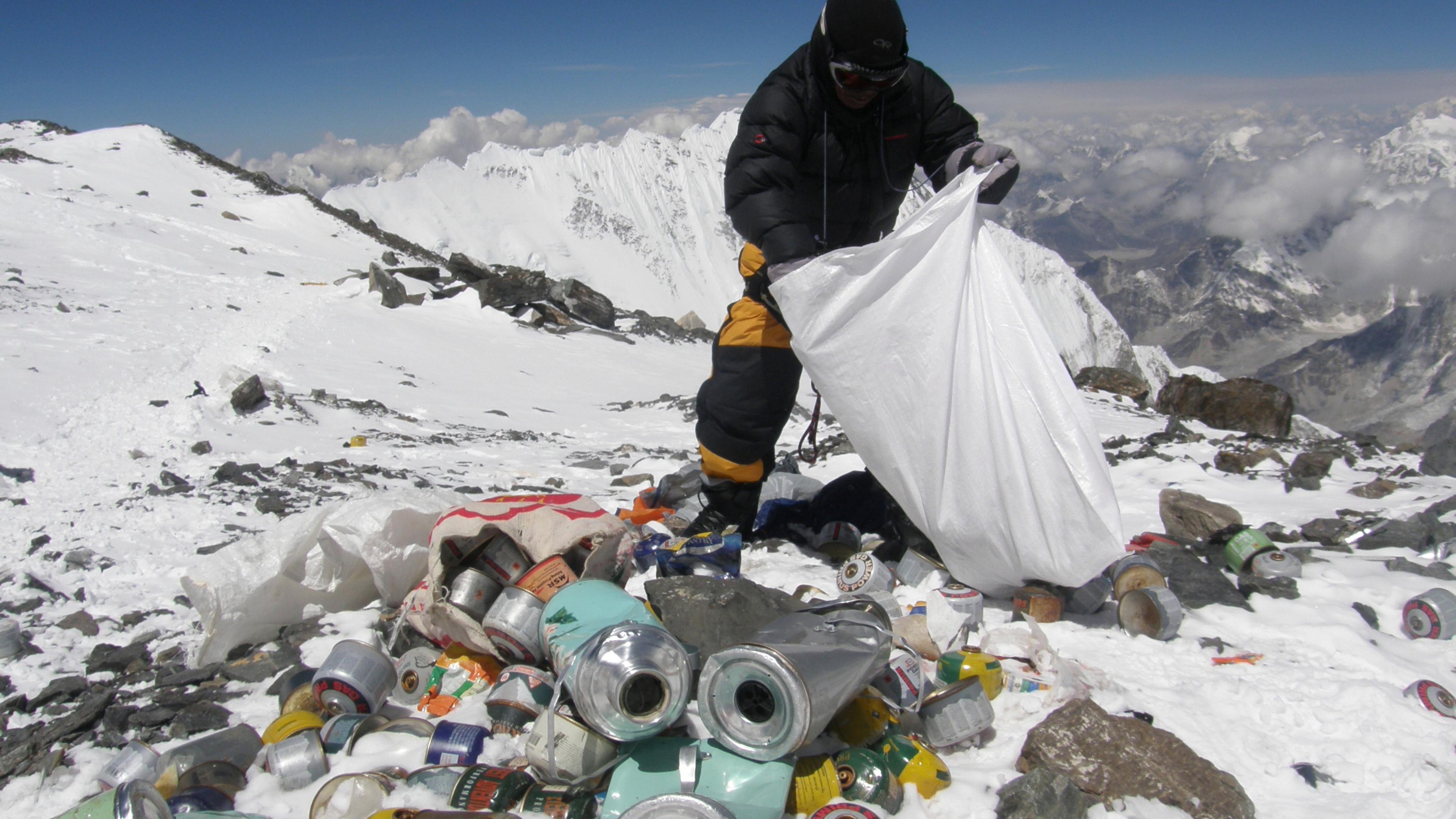 Photo of a person collecting rubbish, including cans, on a snowy mountain peak with a mountainous landscape in the background.