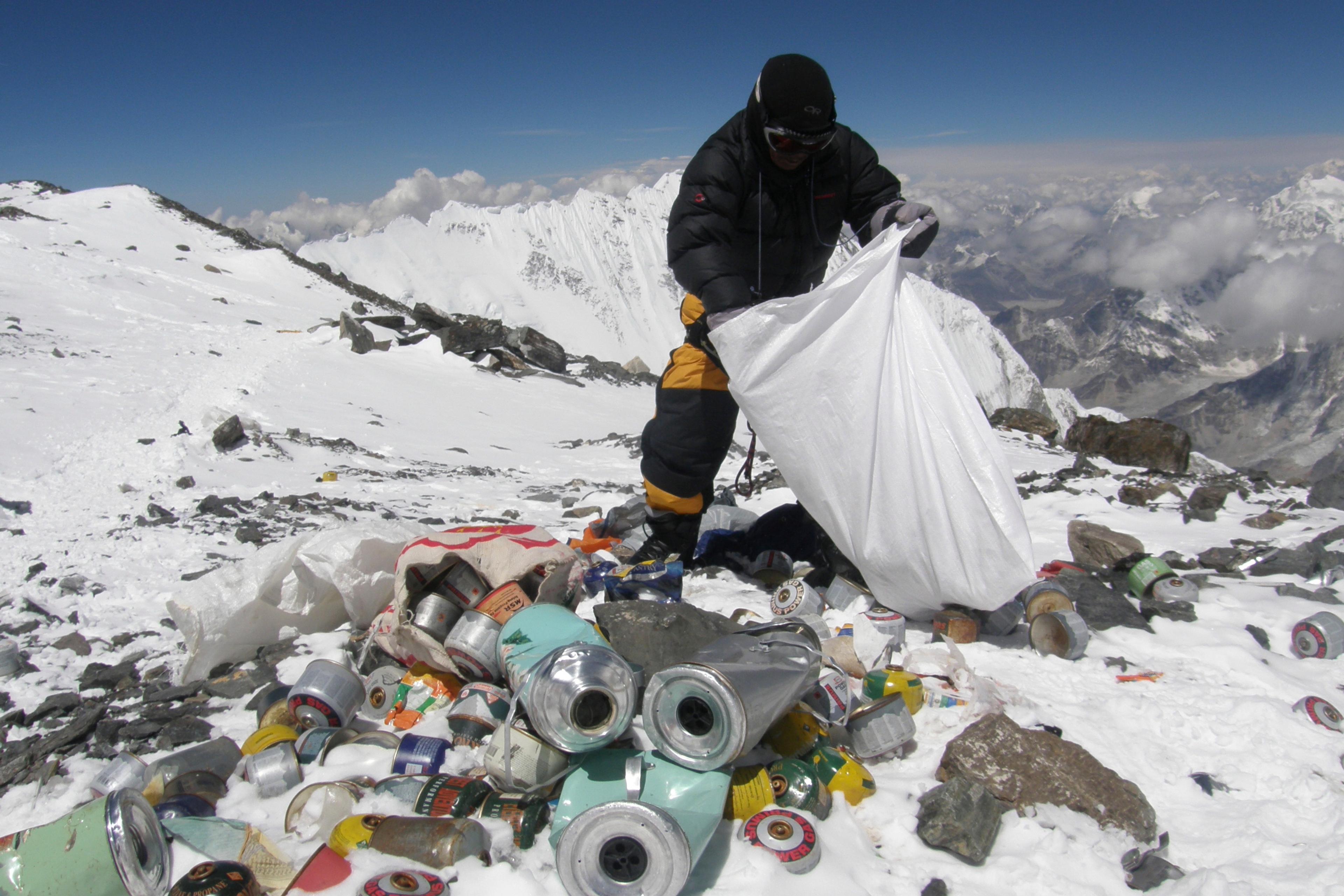 Photo of a person collecting rubbish, including cans, on a snowy mountain peak with a mountainous landscape in the background.