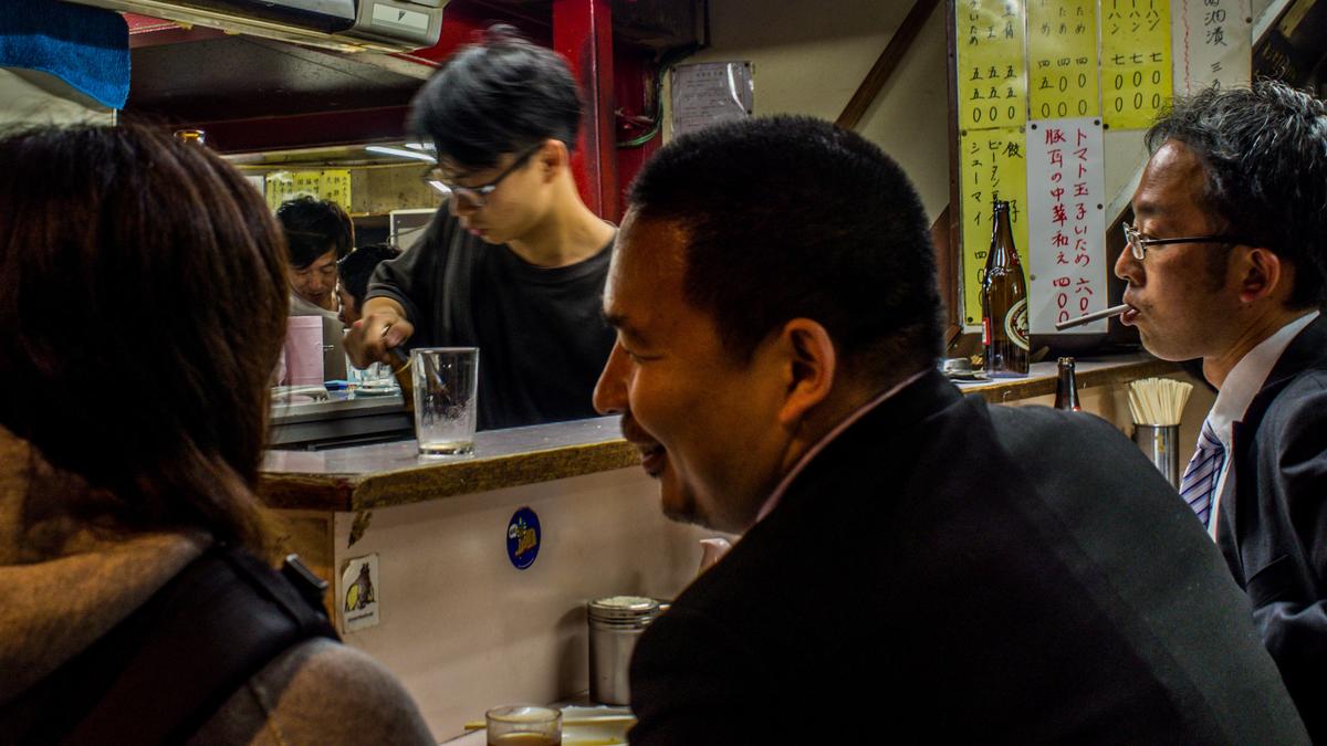 People in a small, well-lit Japanese restaurant or bar, with a menu on the wall and beverages on the counter.