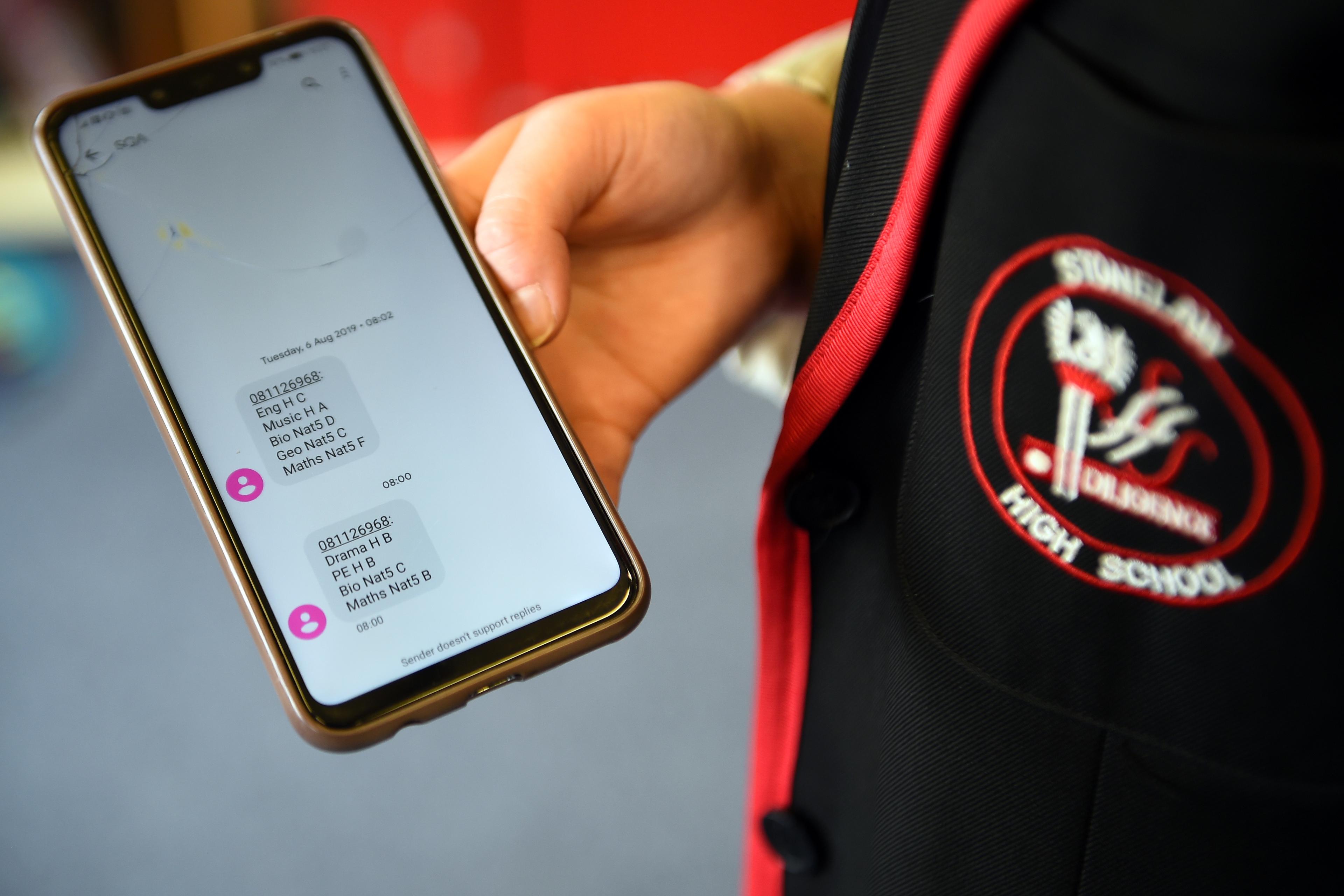 Photo of a person holding a phone showing messages with exam results next to a school uniform badge.