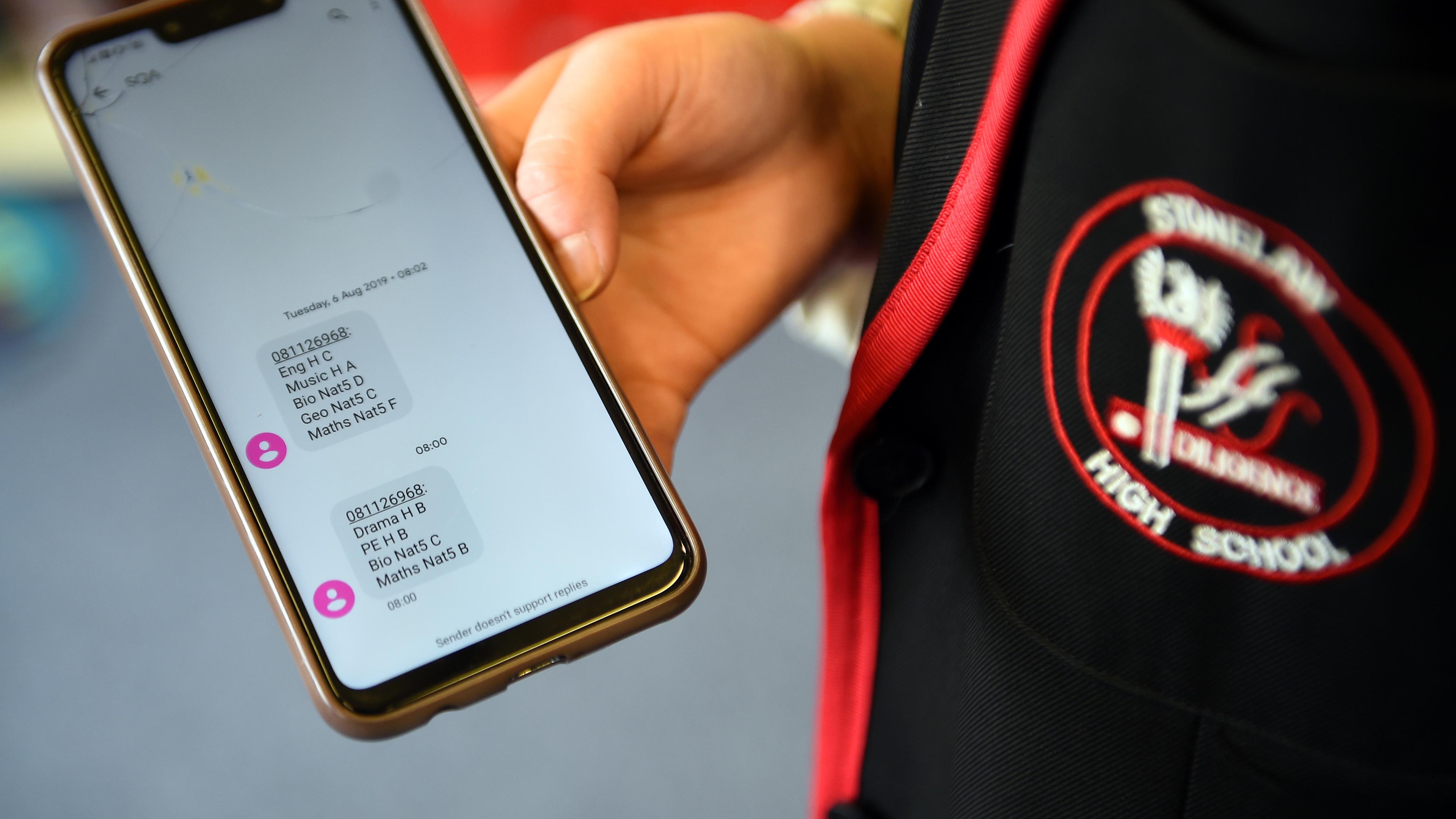 Photo of a person holding a phone showing messages with exam results next to a school uniform badge.