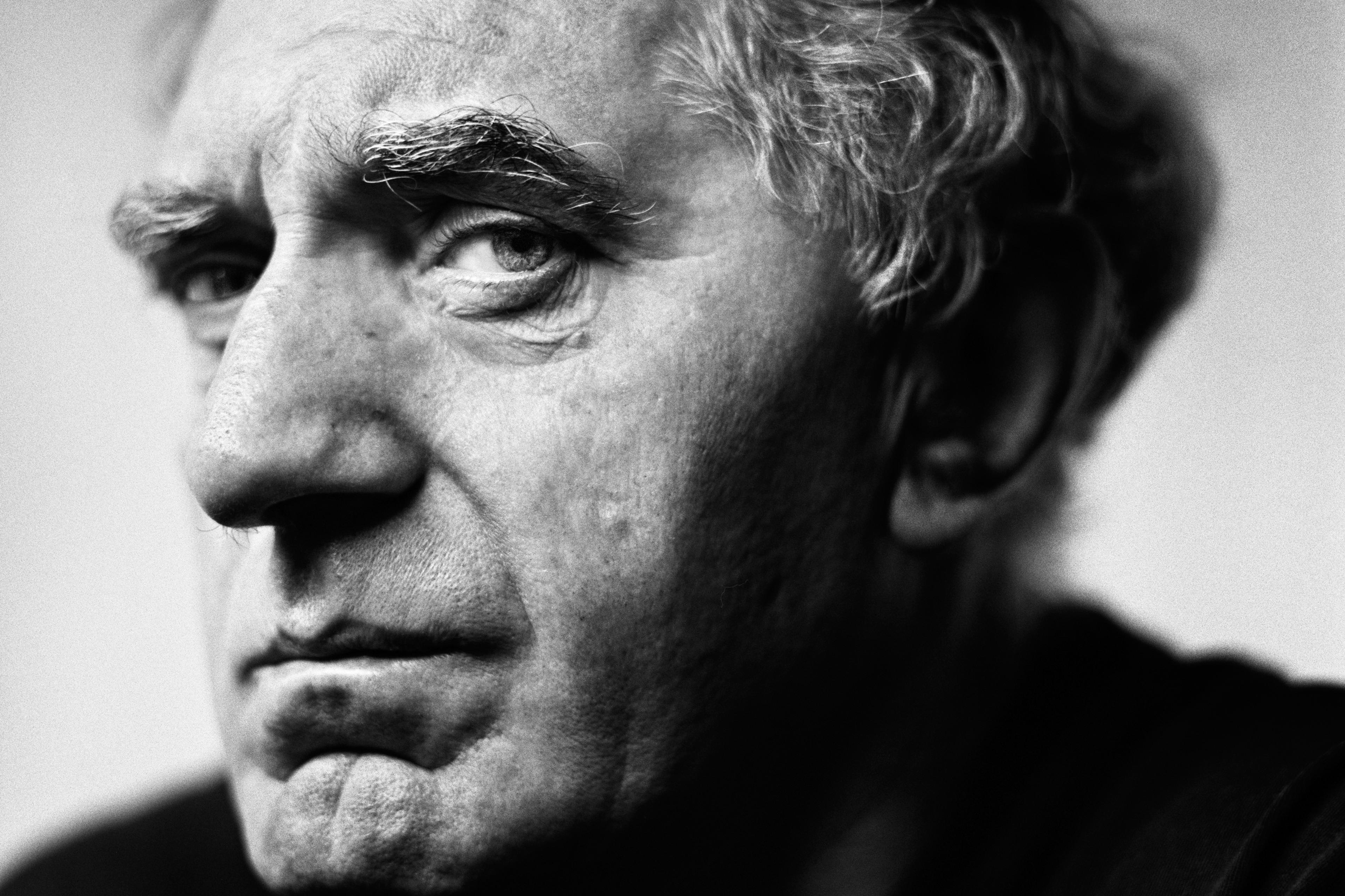 Black and white portrait photo of an older man with curly hair and deep-set eyes, looking intently to the left.