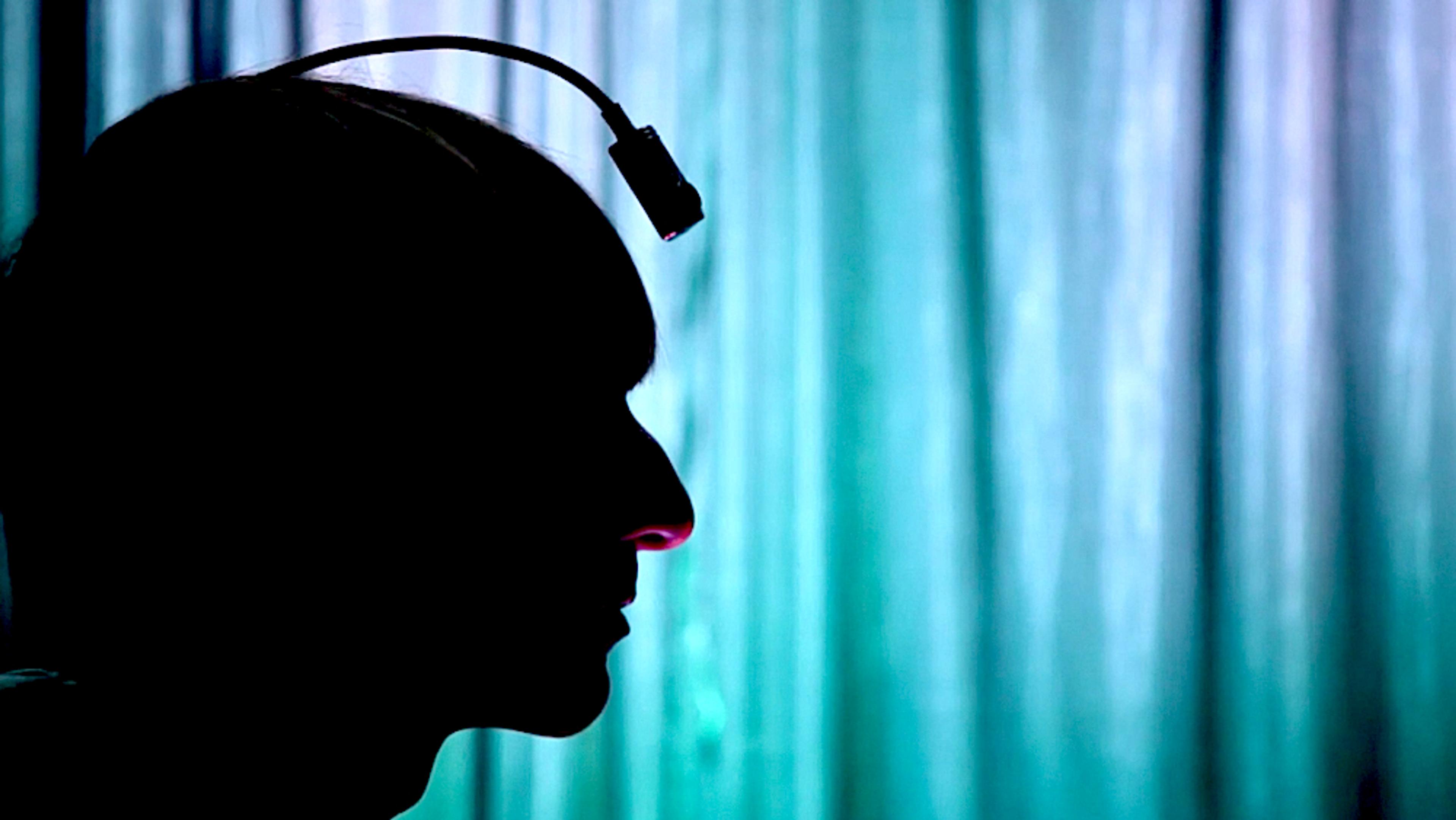 Silhouette photo of a person with a device attached to their head against a background of vertical blue-green curtains.