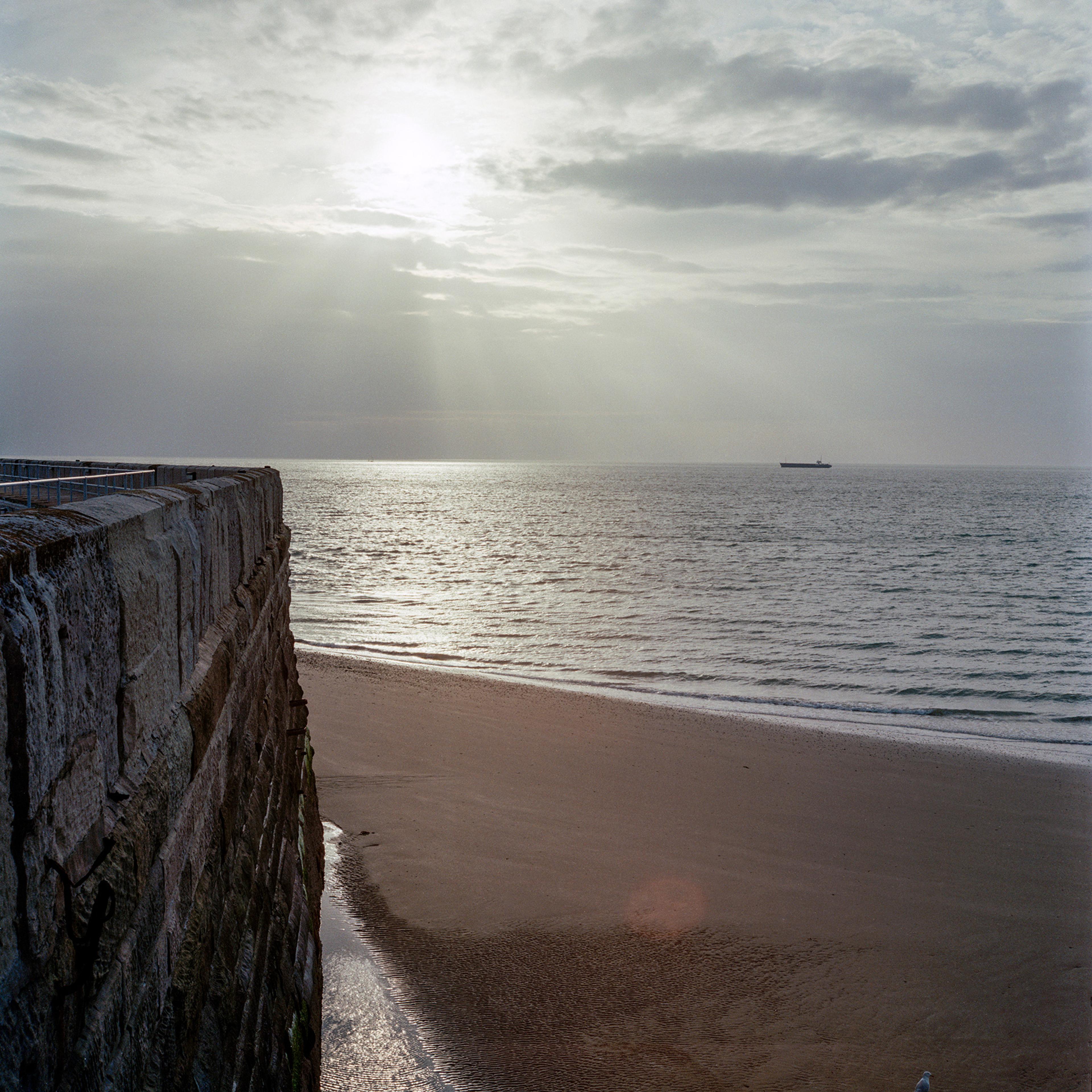 A coastal scene with a stone wall on the left, sandy beach below, and calm sea extending to the horizon. The sun is partially obscured by clouds, casting rays over the water. A distant ship is visible on the horizon.