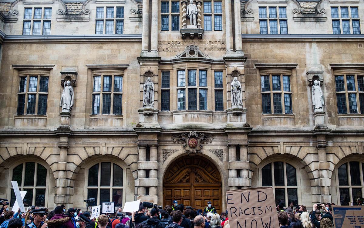 Photo of a protest in front of a historic building with statues. Signs read “End Racism Now!” and other messages.