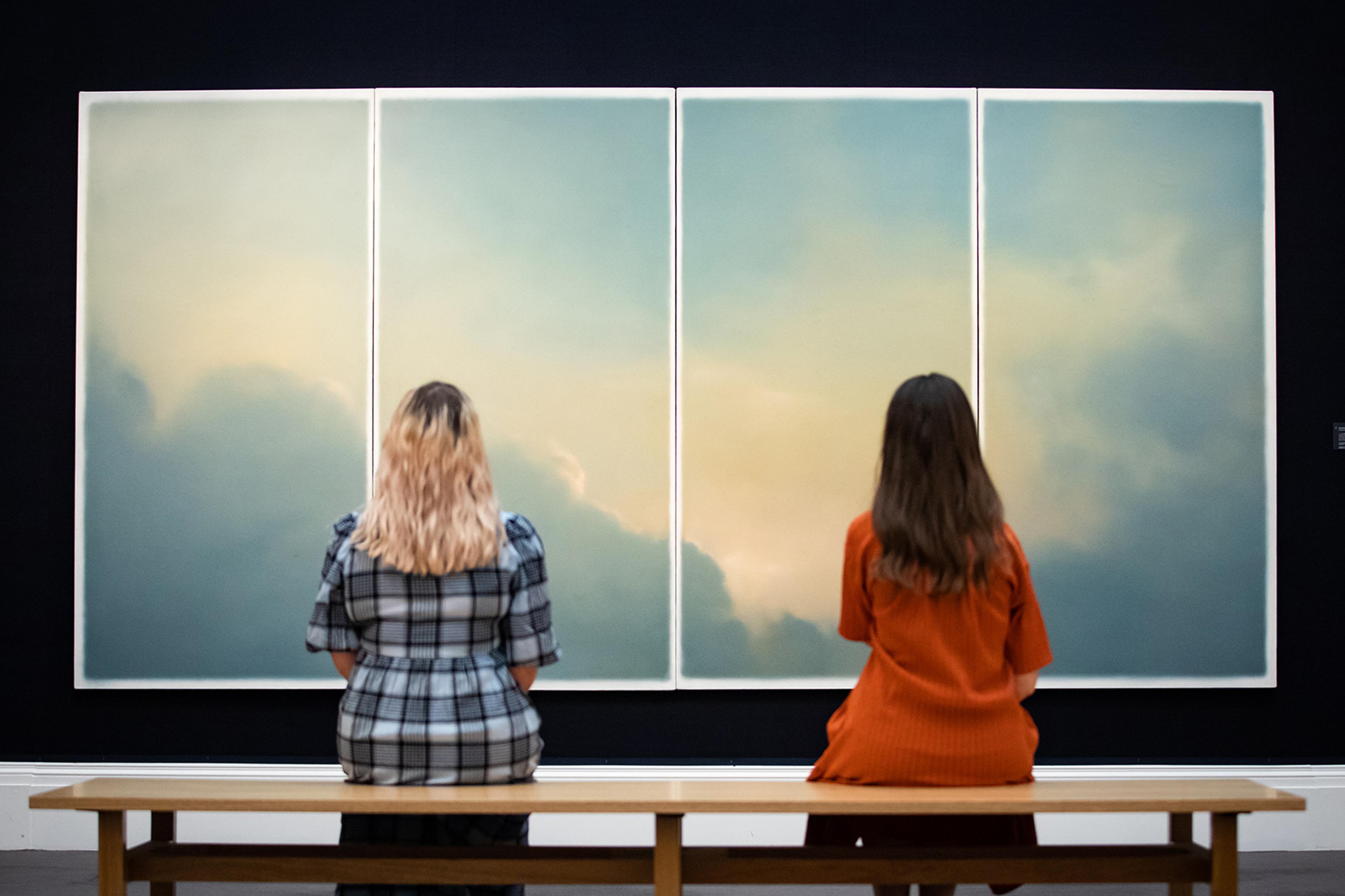 Two women seen from behind seated on a bench viewing a large four-panel painting of a cloudy sky in an art gallery.