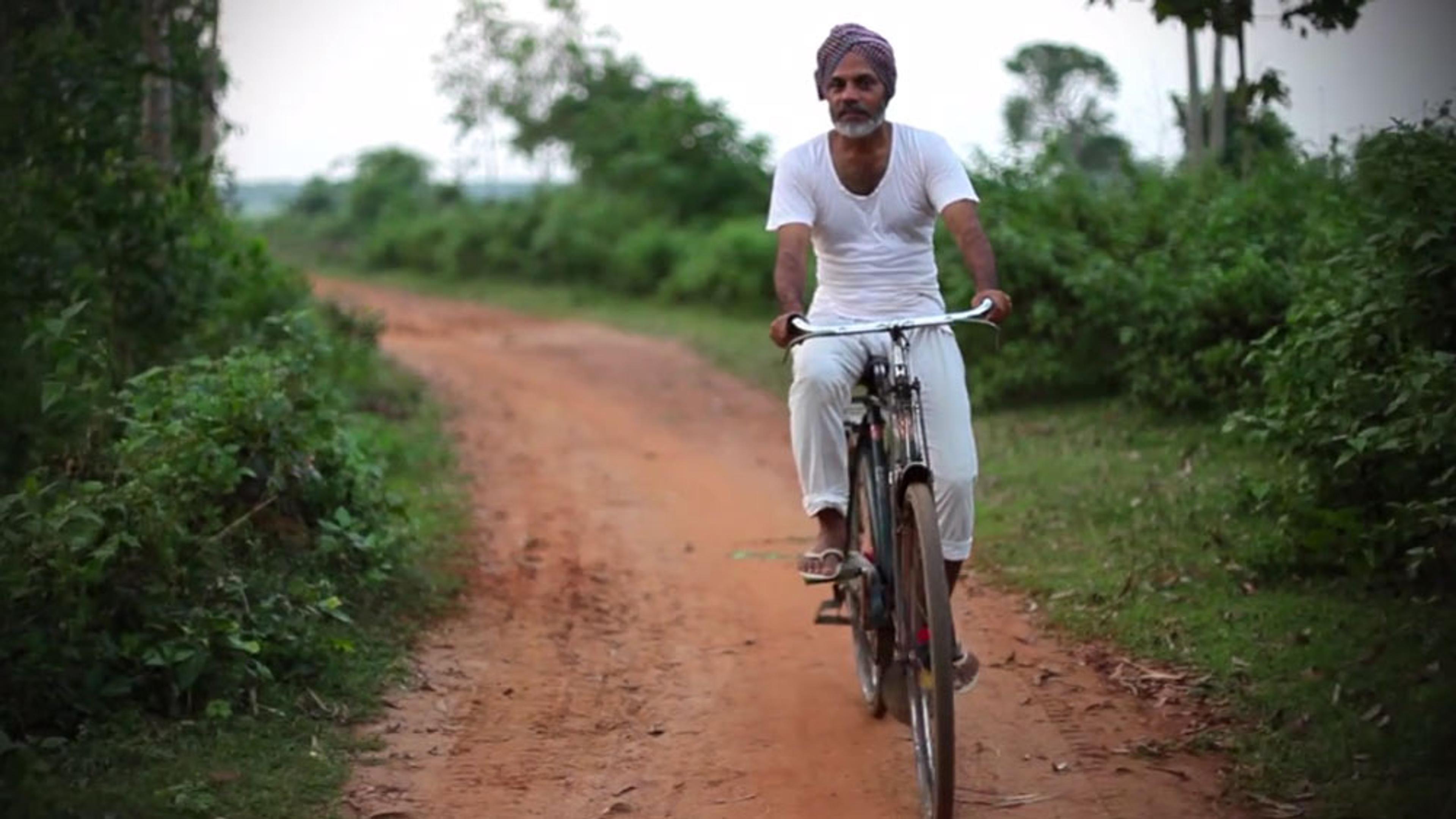 A man wearing a turban and white clothes cycling on a red dirt pathway lined with green foliage and trees in a rural area.