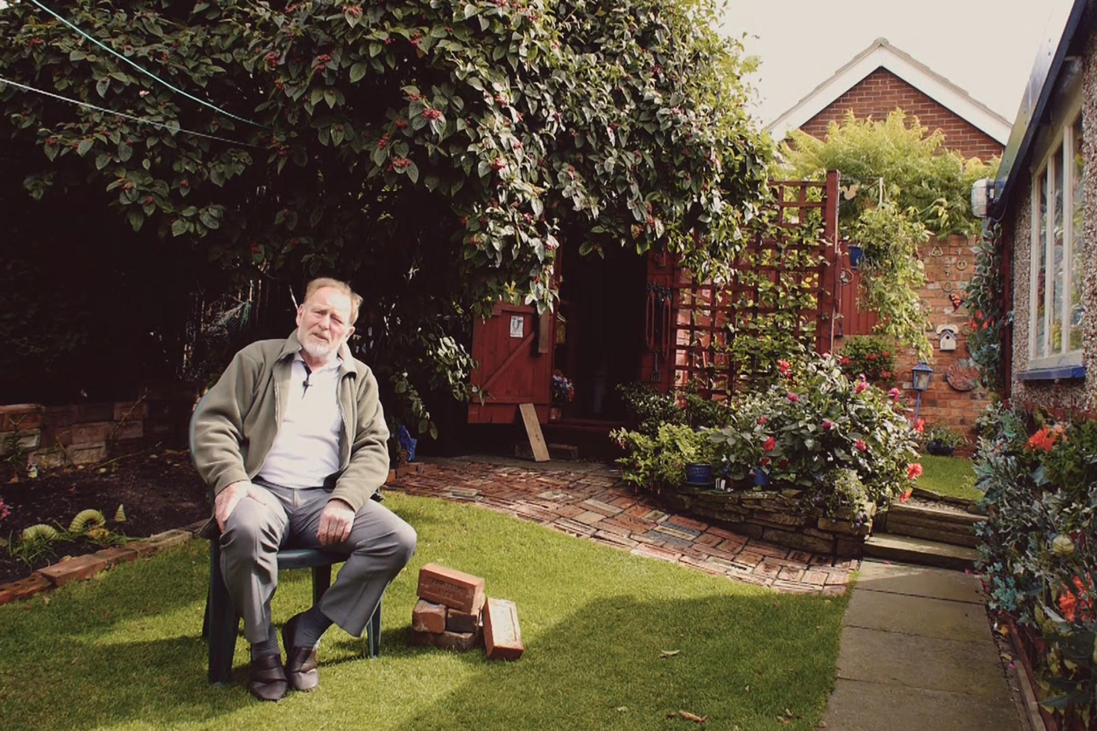 A man sitting on a chair in a lush garden with blooming flowers, a cobbled path, and a red garden shed in the background.