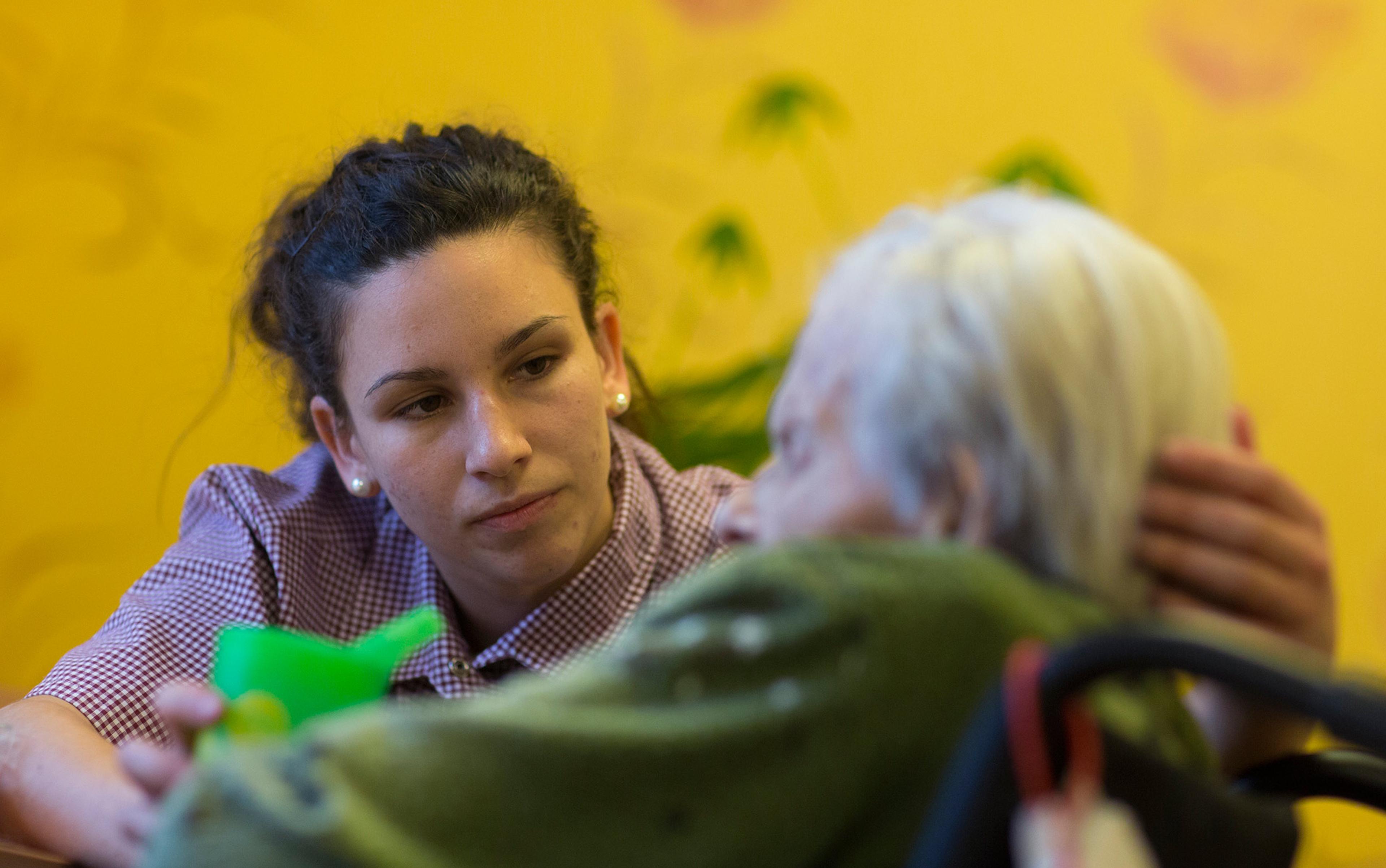 A young woman in a checked shirt caring for an elderly person with white hair, holding their head and a green cup.