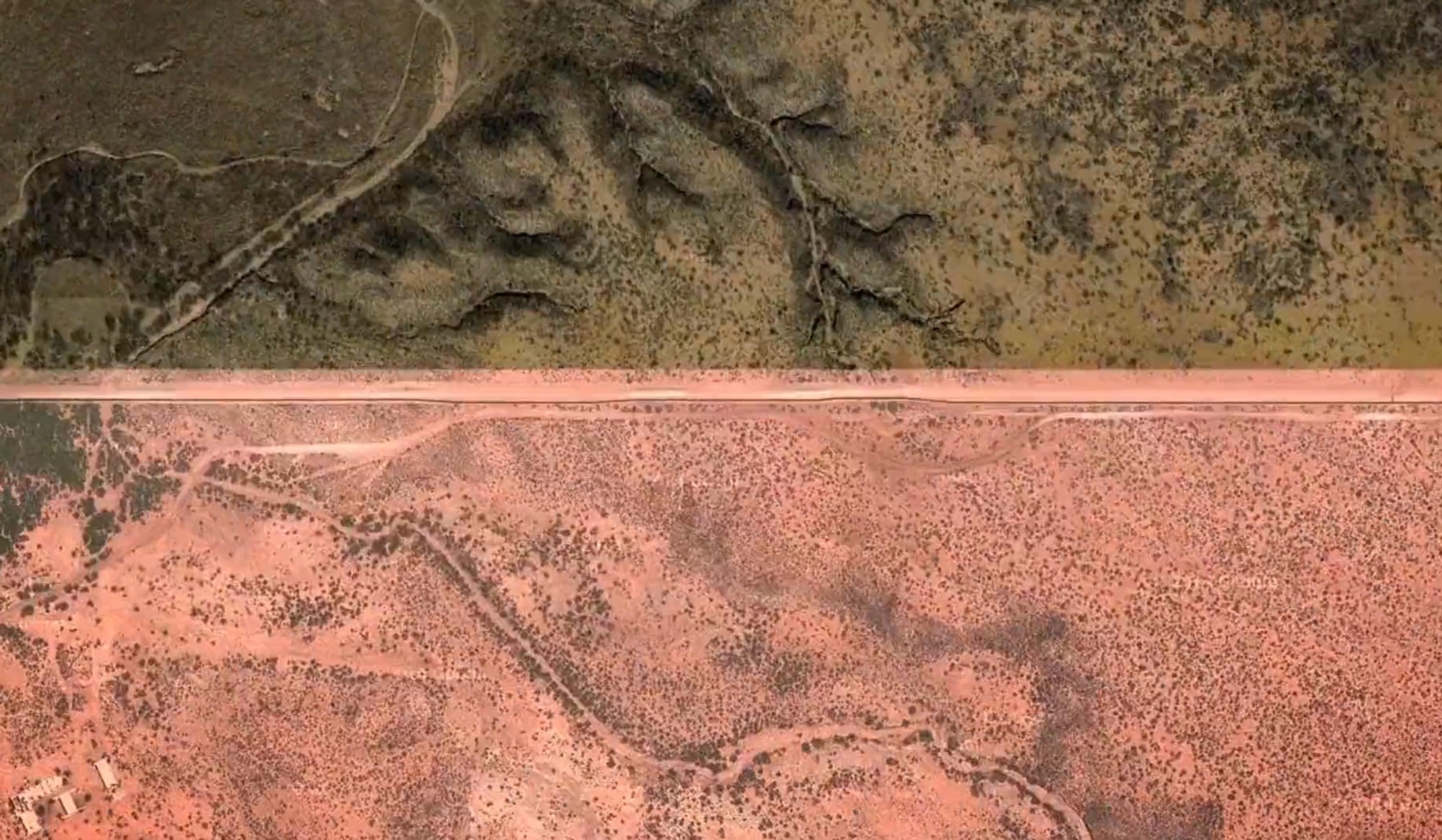 Satellite photo of arid terrain featuring a road that separates darker, more vegetated land from lighter, less vegetated land.