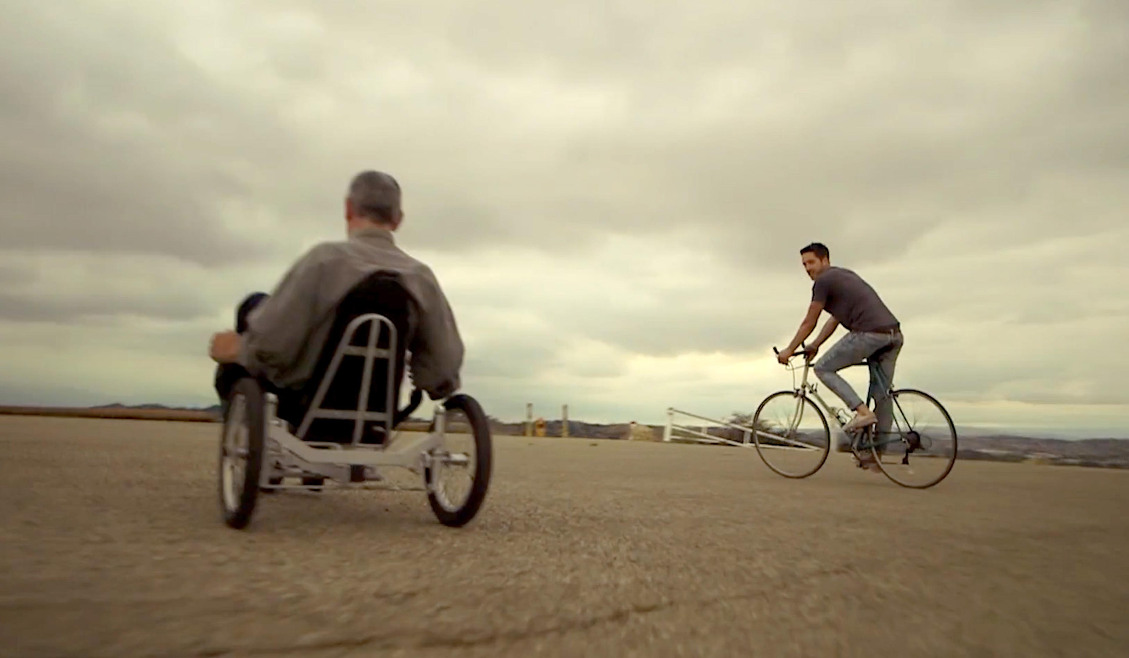 Man on recumbent bicycle and another on road bike cycling outdoors with cloudy sky in the background.