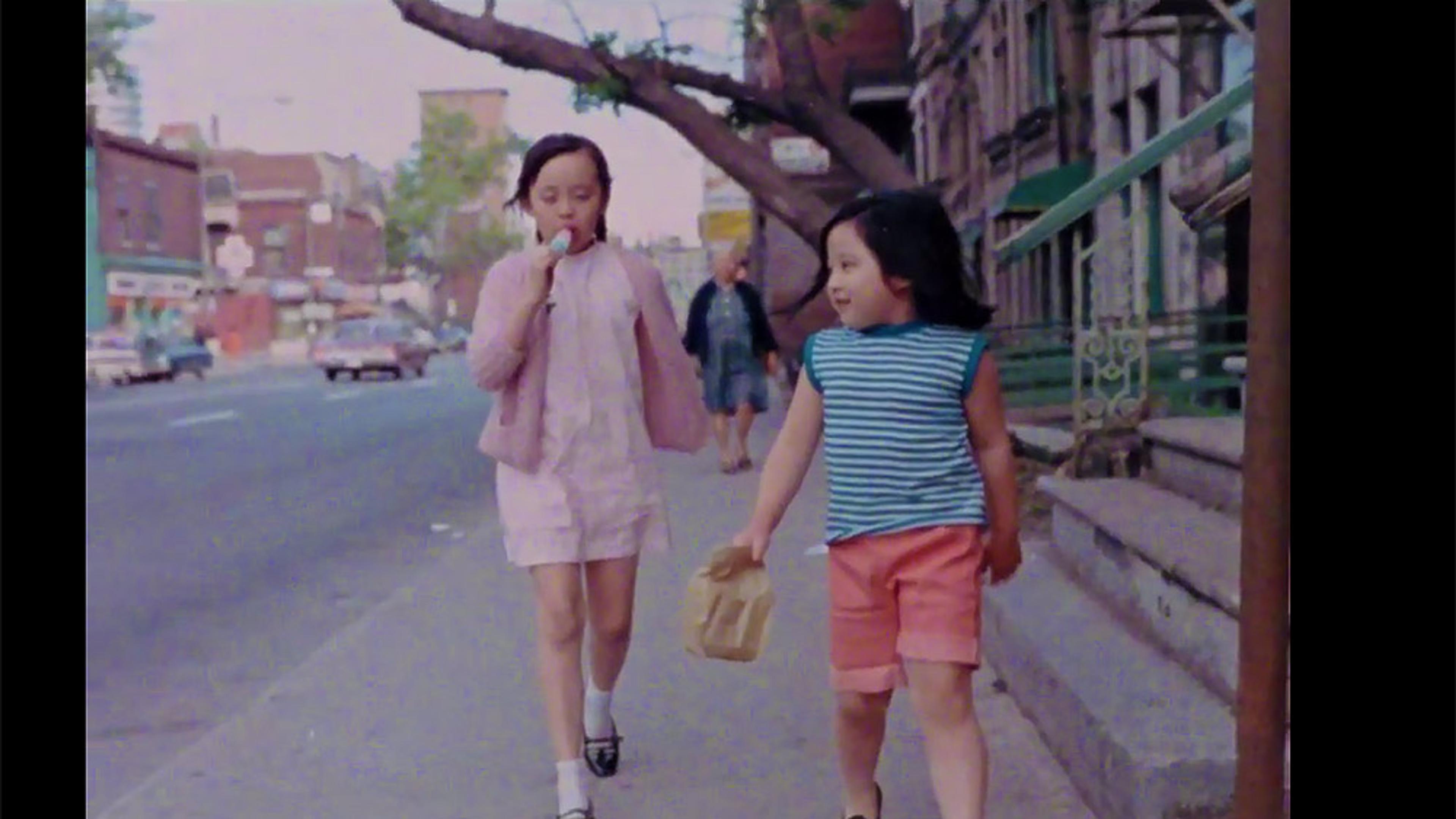 Photo of two young girls walking on a city street; one eating a lollipop, the other holding a paper bag, buildings in the background.