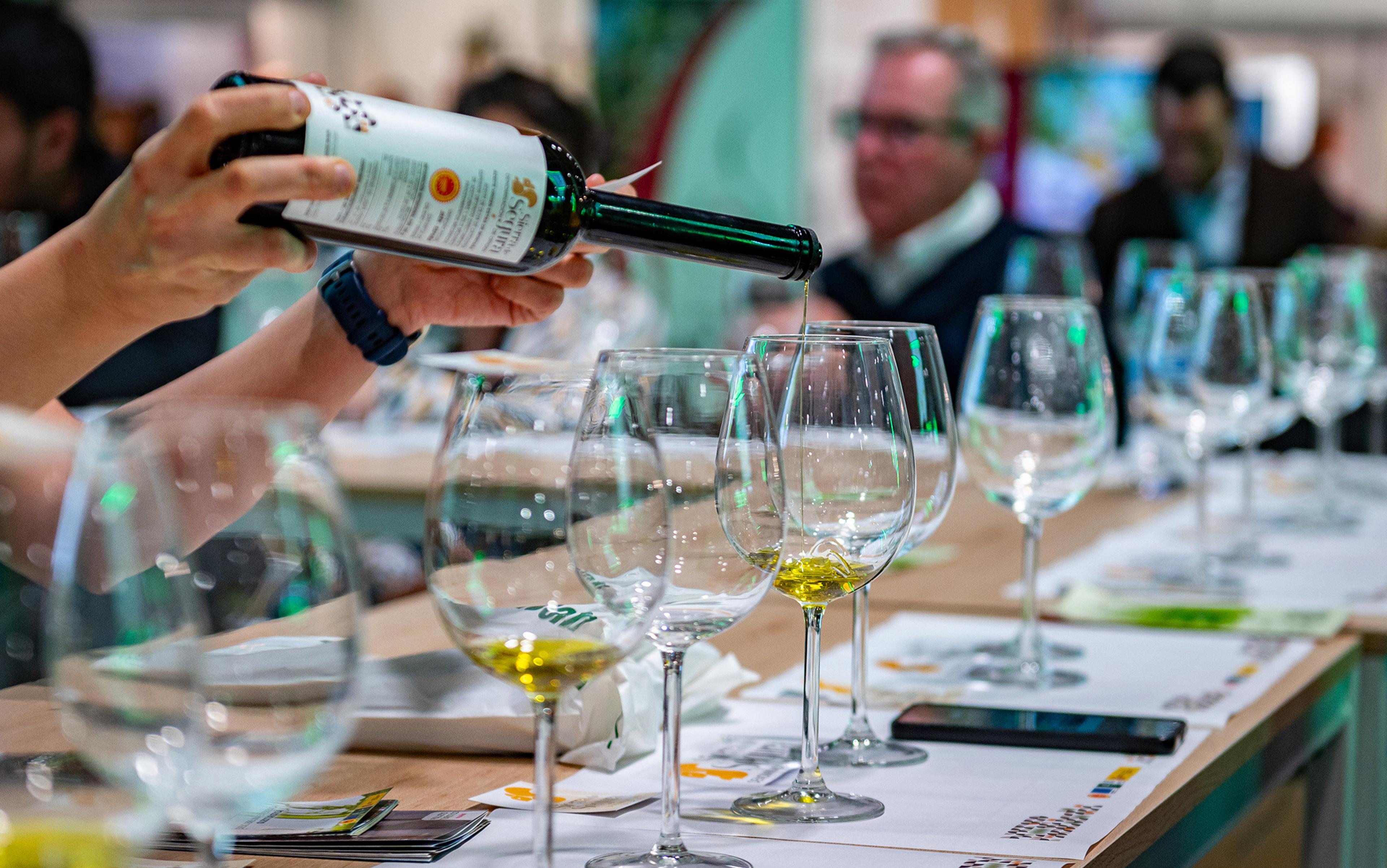 A person’s hands pouring olive oil into a row of wine glasses with people blurred in the background at a tasting event.