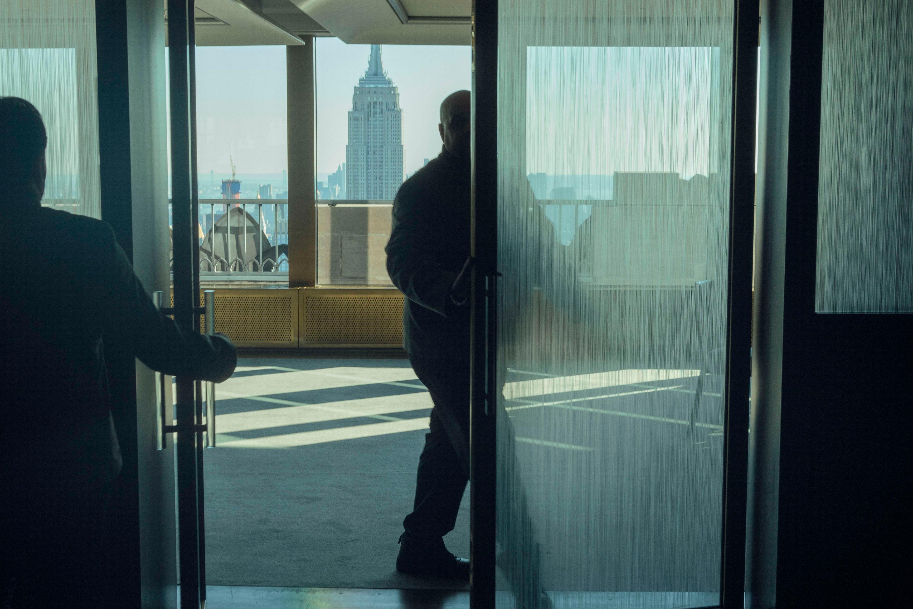 Photo of two people entering a room with a view of a cityscape through large glass doors, Empire State Building visible.