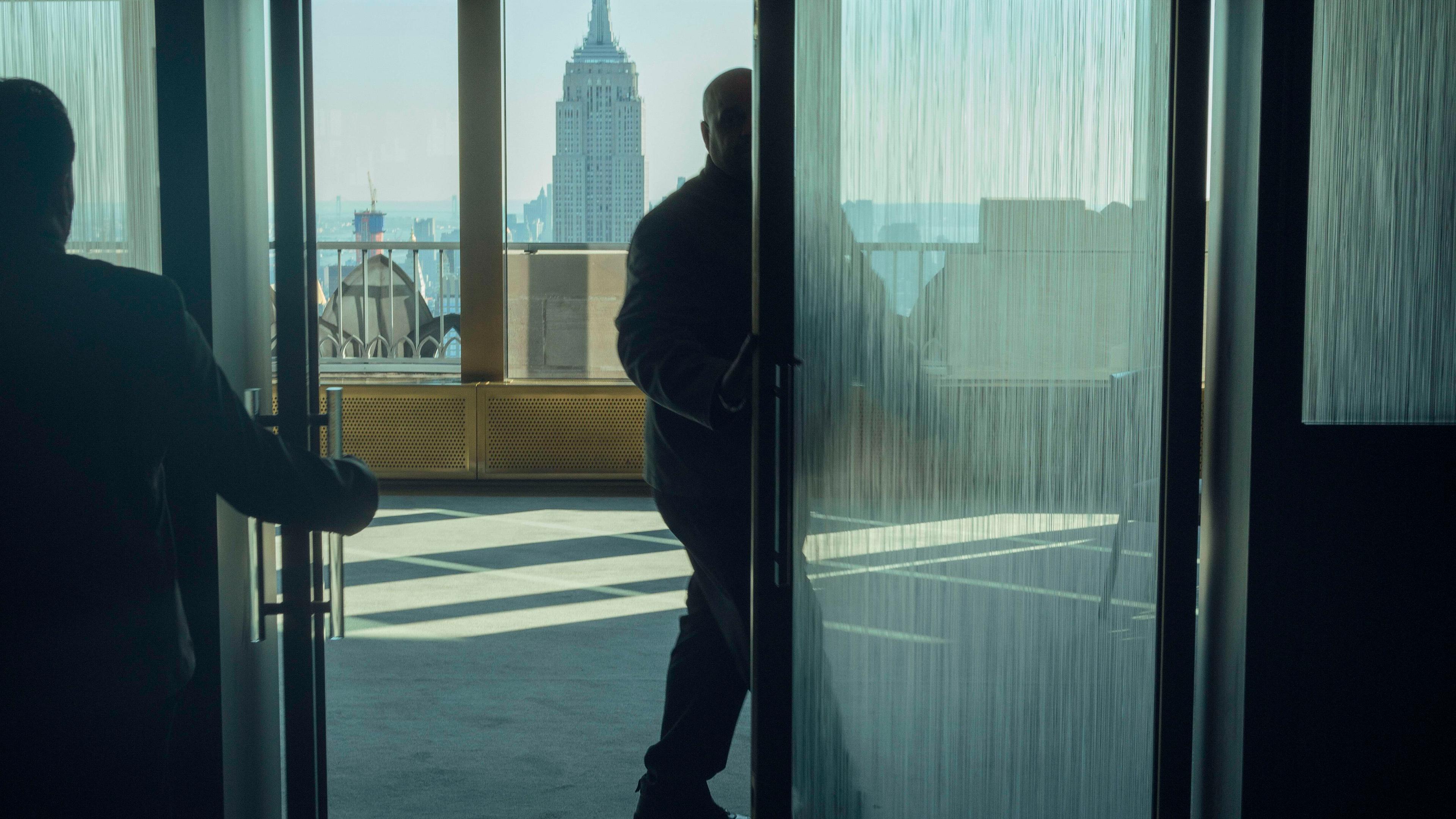 Photo of two people entering a room with a view of a cityscape through large glass doors, Empire State Building visible.